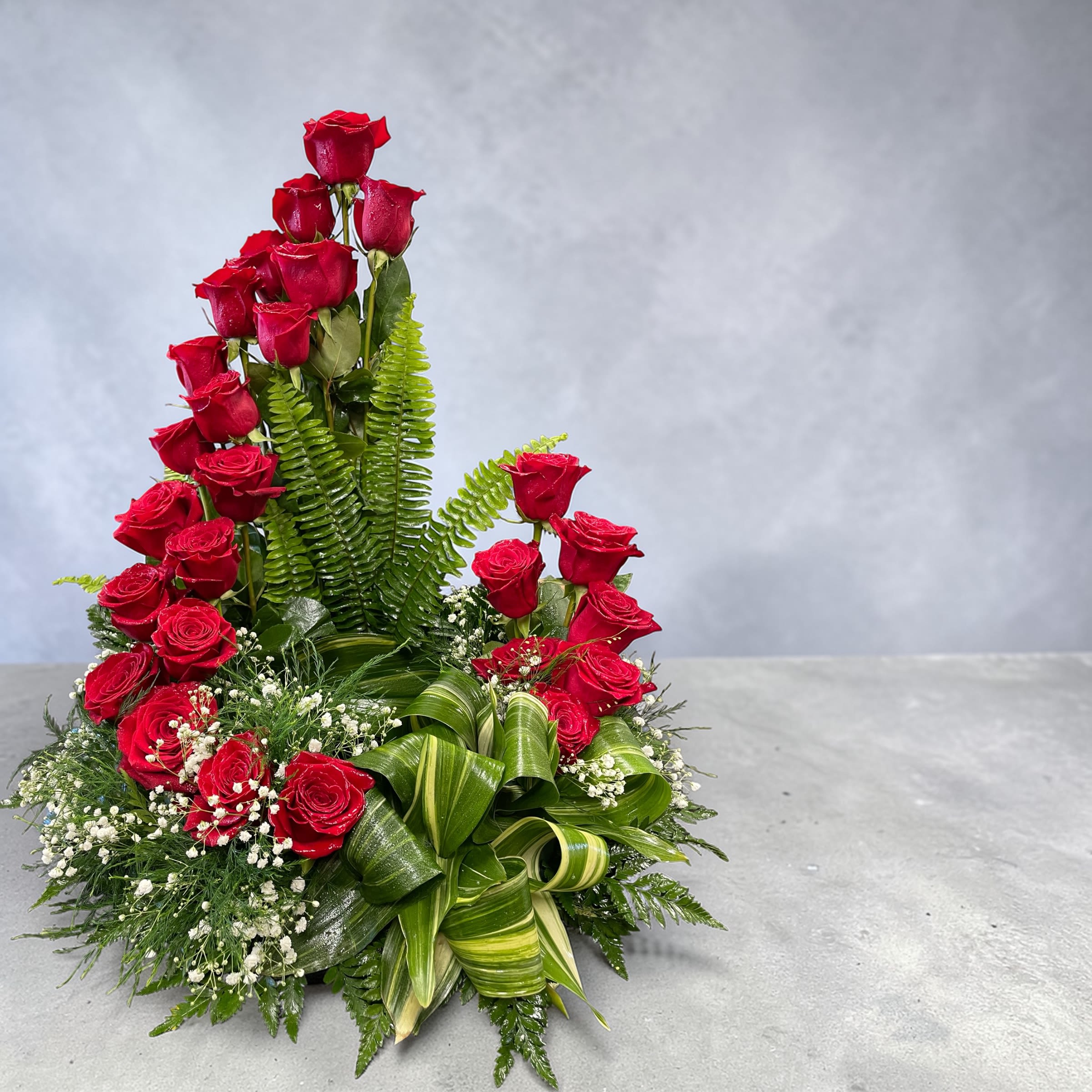 Red Rose Spiral Cremation Wreath  - A dramatic cascade of vivid red roses rises in a sculptural triangular spiral, creating a strong vertical focal point for a sympathy and funeral flower arrangement. This red rose funeral arrangement is designed to honor a loved one with the timeless language of roses-its bold color and formal silhouette convey deep love and reverence so the person you're thinking of feels remembered and cherished.  Multiple tiers of tightly set red roses climb from a low round container, framed by lush fern fronds and broad variegated leaf loops that give the piece architectural structure. Small clusters of white filler blooms at the base soften the composition and add gentle contrast to the deep greens and reds, while the low container keeps the presentation neat and suitable for placement near an urn or at a memorial service.  Appropriate for family members, partners, or anyone who found special meaning in classic romance, this arrangement reads as a heartfelt tribute that also works for anniversary memorials or other intimate remembrance moments. Arrives ready to display in the included low round container; mist the blooms lightly and keep away from direct heat to extend freshness.