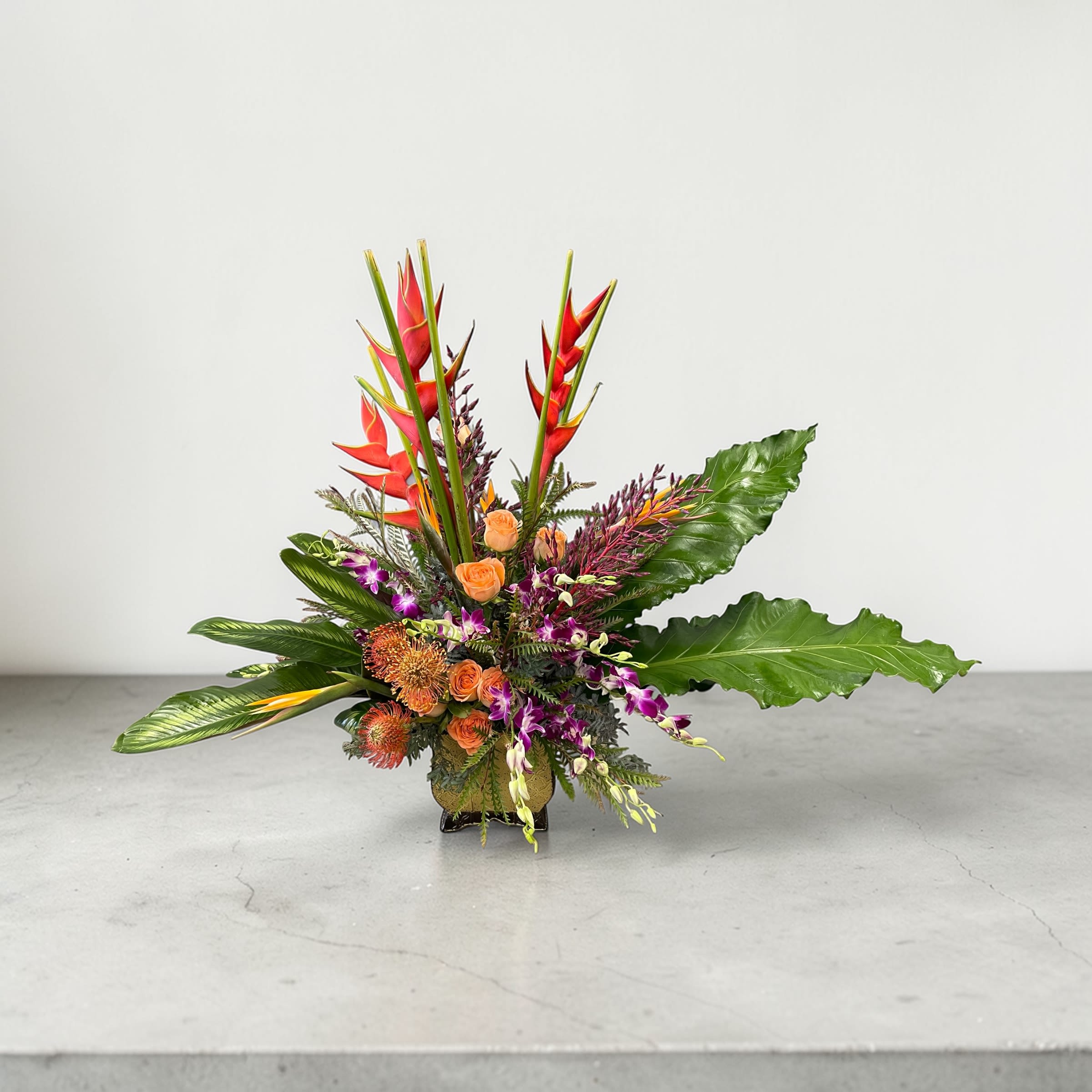 Tropical arrangement with orange roses, protea, purple orchids, and red heliconia in a low glass vase