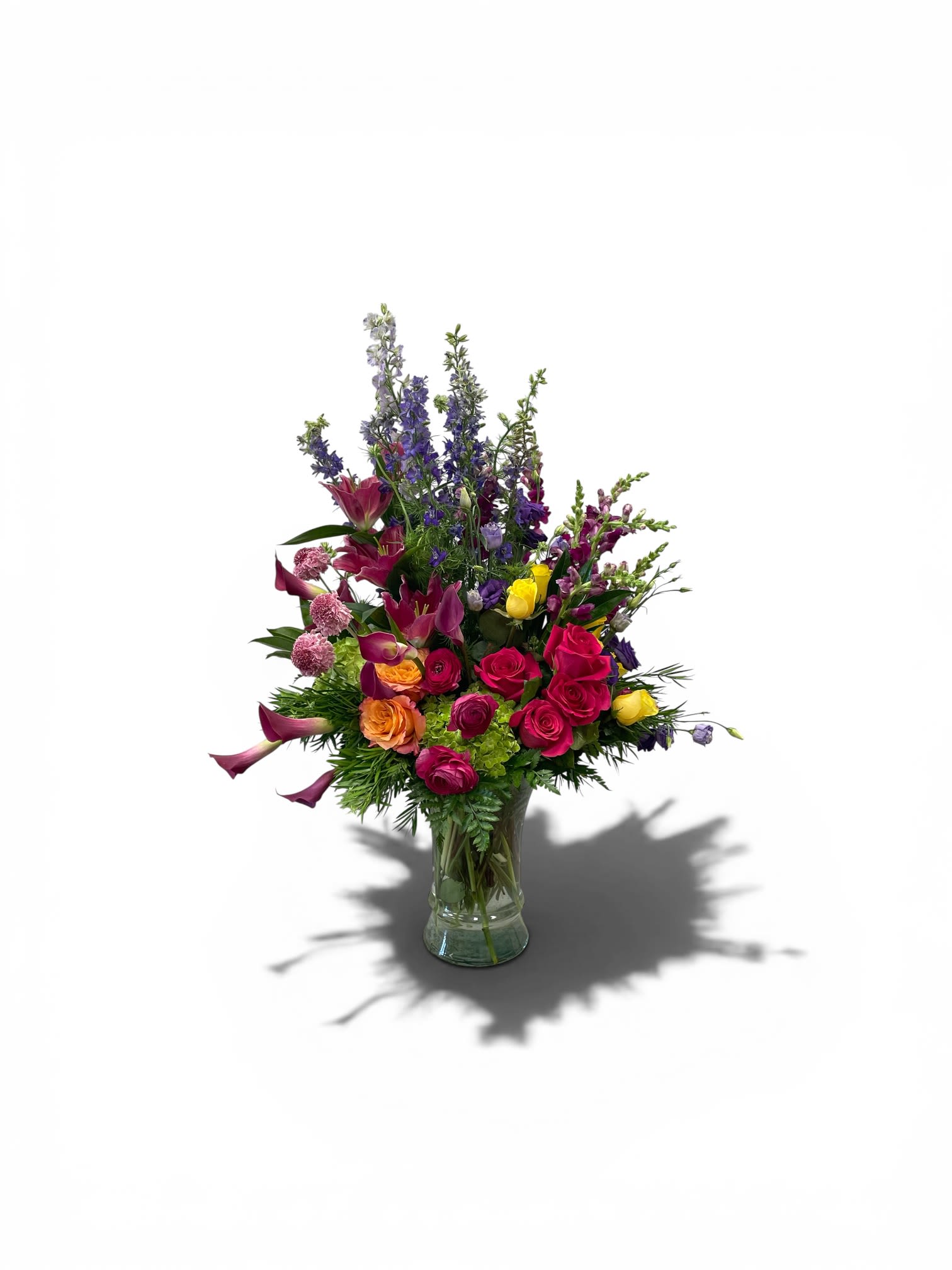 Vibrant Romance  - A bold cascade of color greets the eye - hot pink roses and deep burgundy mini calla lilies punctuated by bright yellow and orange blooms create the heart of this flower arrangement, perfect as an anniversary roses bouquet, a birthday surprise, or a vibrant Mother's Day gift. Spikes of purple and blue delphinium and snapdragons rise above the cluster to give the piece a tall, garden-like silhouette that reads both celebratory and romantic; it's the kind of arrangement that makes the person you're thinking of feel truly seen and adored.  Set in a clear glass vase, the design layers lime-green accent blooms and textural filler throughout for contrast and visual depth, with soft touches of lavender, peach and white balancing the saturated palette. The mix of roses, mini calla lilies, delphinium, snapdragons and hydrangea-like fullness gives varied texture and lasting presence on a mantel, dining table, or bedside. Ideal for occasions from graduations and housewarmings to get well wishes, sympathy, or just because moments, this arrangement also suits seasonal celebrations like Easter and springtime events while remaining a confident gift any day of the year. Care: refresh water every two days and trim stems slightly when re-cutting to extend vase life.