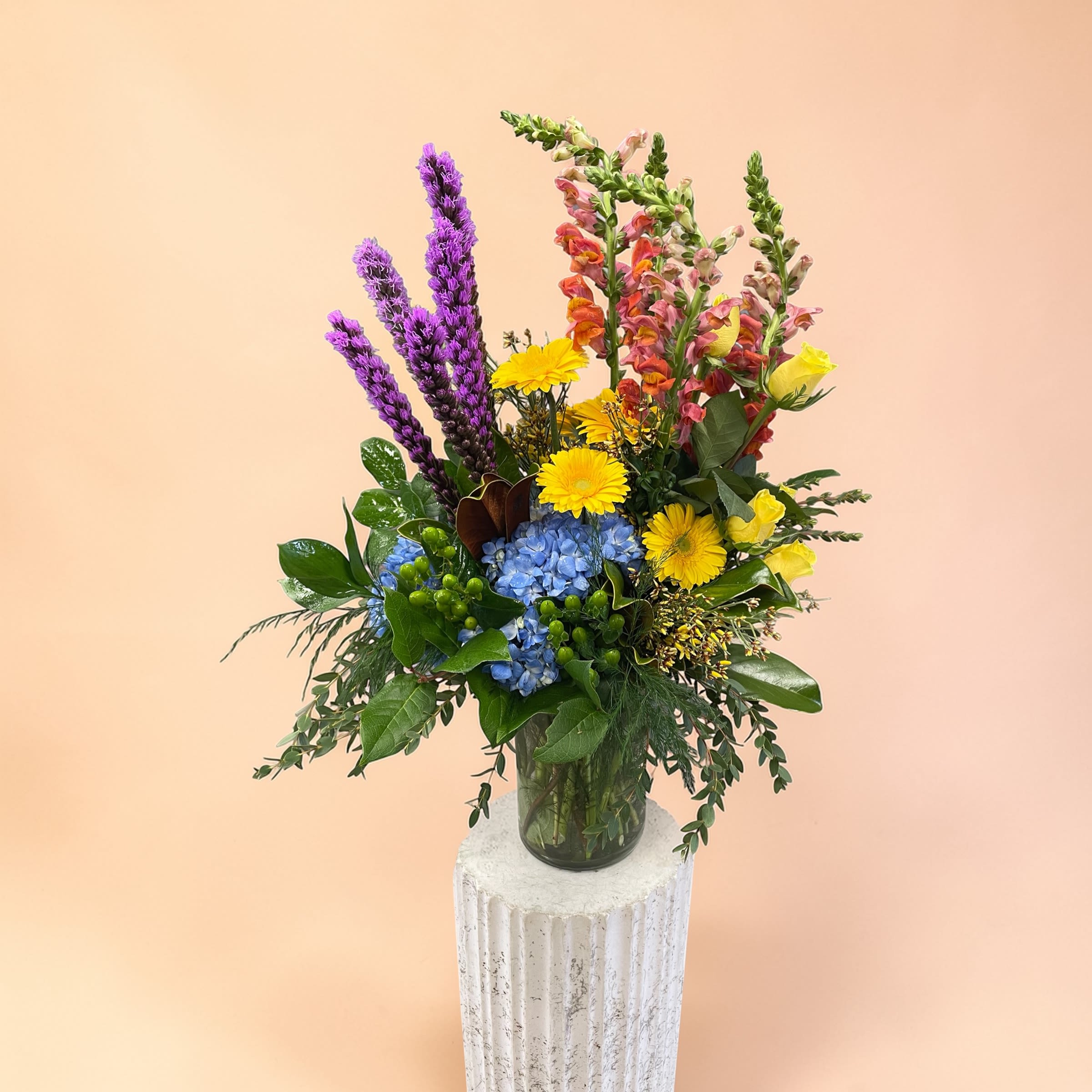 Beauty and Solace  - A tall, airy arrangement that feels like a sunlit garden in a clear glass vase - perfect as a birthday bouquet, Mother's Day surprise, or a cheerful get well gift. This flower arrangement for spring and Easter occasions lifts the eye with bright yellow daisy-style blooms and multicolored snapdragons that create lively vertical accents, while the rounded base brings balance and a casually elegant silhouette.  Blue hydrangeas form the lush foundation, contrasted by pinks, purples, peaches, whites and touches of orange and lavender for a playful, mixed-color palette. Textural variety from gerbera daisies and snapdragons gives the piece movement and depth; the flowers arrive arranged in a clear glass vase and presented on a white textured pedestal stand, making it ready to display on a mantel, entry table, or as a centerpiece. Sized to make a visible impact without overwhelming a room, it's an uplifting choice for graduations, anniversaries, housewarmings, thank-you sentiments, sympathy, or simply to say "thinking of you."  This composition is designed to brighten someone's day - she'll feel genuinely celebrated and seen when these joyful colors arrive. For best longevity, keep the vase filled with fresh water and out of direct heat; consider this mixed bouquet for seasonal holidays, everyday occasions like Just Because or Congratulations, and as a versatile option for everything from weddings and proms to sympathy and remembrance.