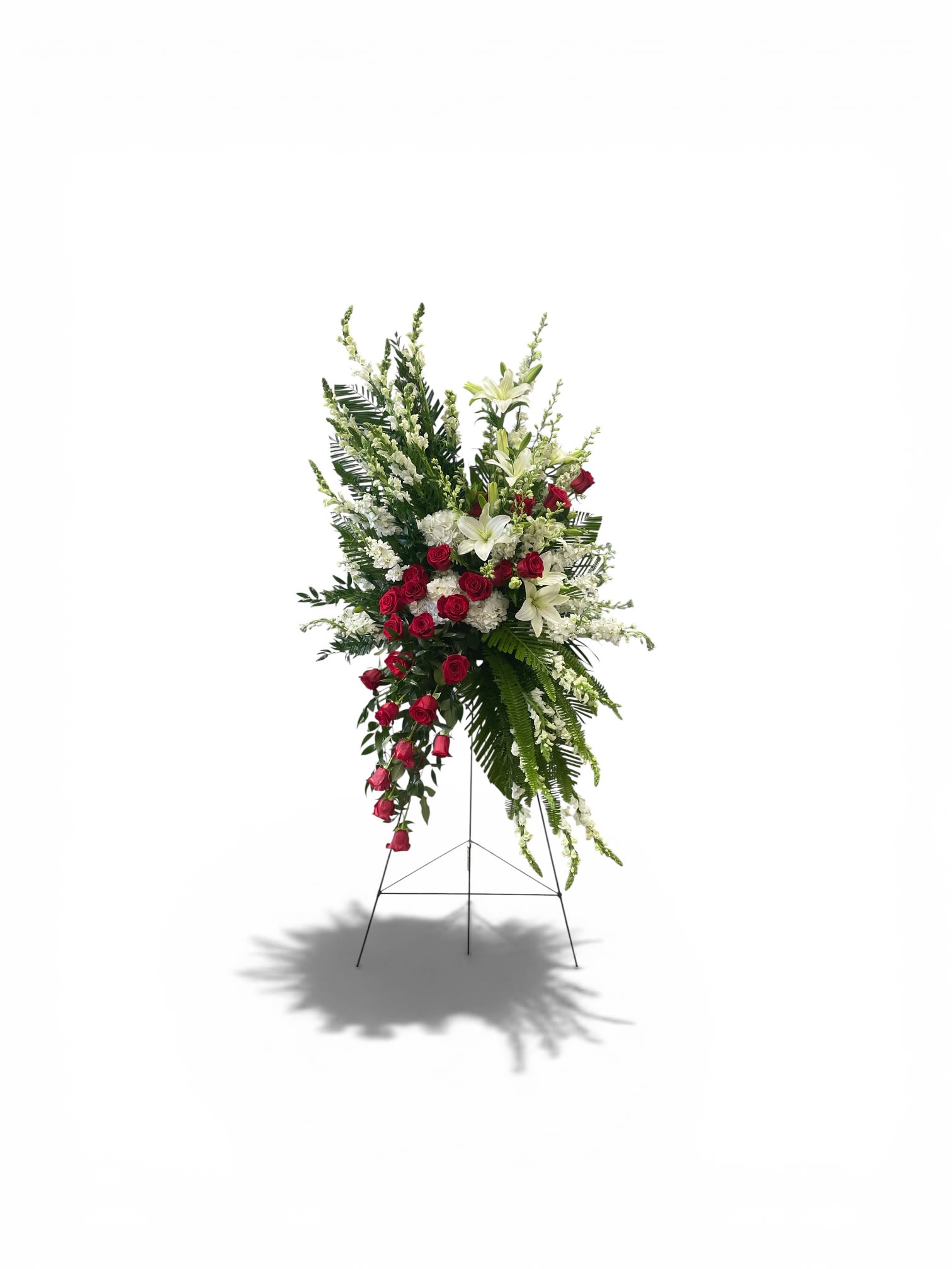 The Modern Bleeding Heart Spray - A striking asymmetric spray of red roses and white lilies rises on a metal easel stand, creating a bold visual statement for sympathy and funeral services. The Modern Bleeding Heart Spray places clustered red roses near the center that cascade downward like a ribbon of deep color, while white lilies and additional white blossoms reach upward and outward to lend height and a serene, formal silhouette-the kind of arrangement that helps the person you're honoring feel deeply remembered.  Carefully arranged snapdragons and chrysanthemums add vertical texture and soft green accents that balance the red-and-white palette and give the spray a structured, ceremonial presence. Mounted on a sturdy metal easel, this standing arrangement is designed for visibility in a chapel or memorial space; its shape and scale read well from a distance and beside a casket or memorial table.  Choose this spray when you want a message of heartfelt respect and lasting remembrance-appropriate for close family tributes or to convey sympathy from a group. Standard sizing fits most service spaces; please contact us for delivery timing or special placement requests so the arrangement arrives fresh and properly displayed.