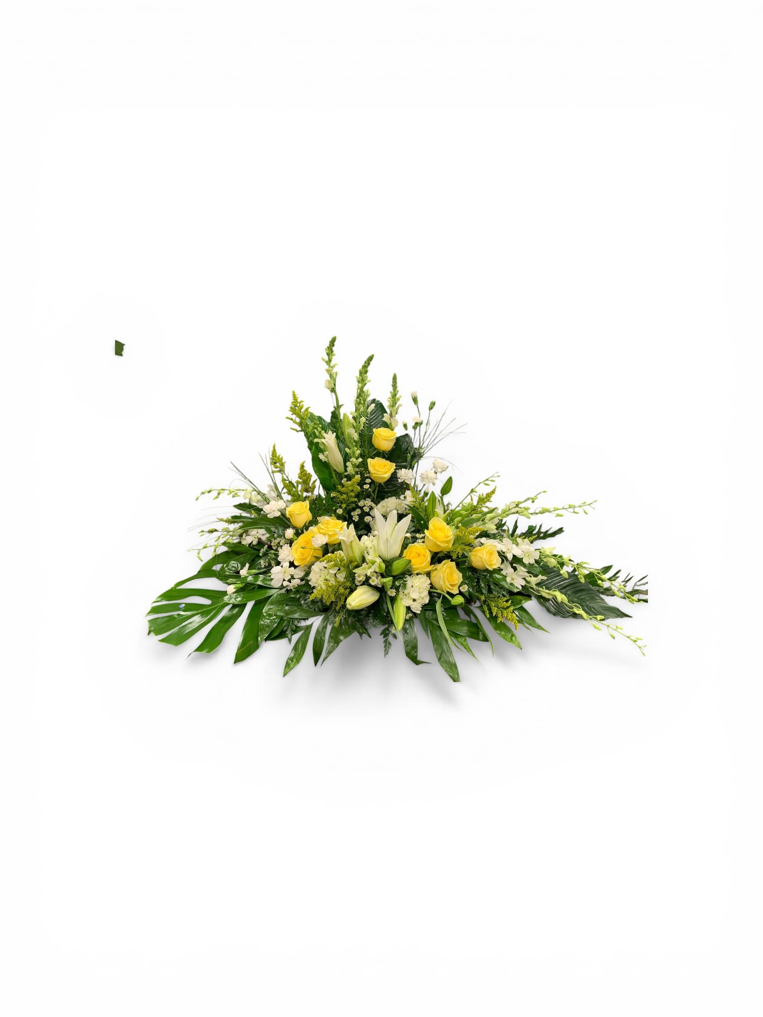 Yellow and White Cremation Wreath  - A low, elongated spray of yellow and white roses and lilies designed for sympathy and funeral settings that still feels quietly uplifting. The horizontal, triangular shape tapers gently at both ends so the arrangement creates a graceful, linear profile on a memorial table - the kind of tribute that helps the person you're honoring feel seen and remembered.  Clusters of bright yellow roses sit beside open and budding white lilies, with other complementary blooms and bold green leaves fanning beneath to add structure and length. The palette of sunny yellow, pure white, and fresh green reads calm and hopeful; the overall size and horizontal format make it ideal as a centerpiece on a low table.  Made to comfort and celebrate a life, this wreath sends a thoughtful message of remembrance and warmth for funeral services or sympathy visits. Arrange for delivery to the service location and keep stems in clean water until placement; if you need a different size or personalization, let us know and we'll help create the right tribute.