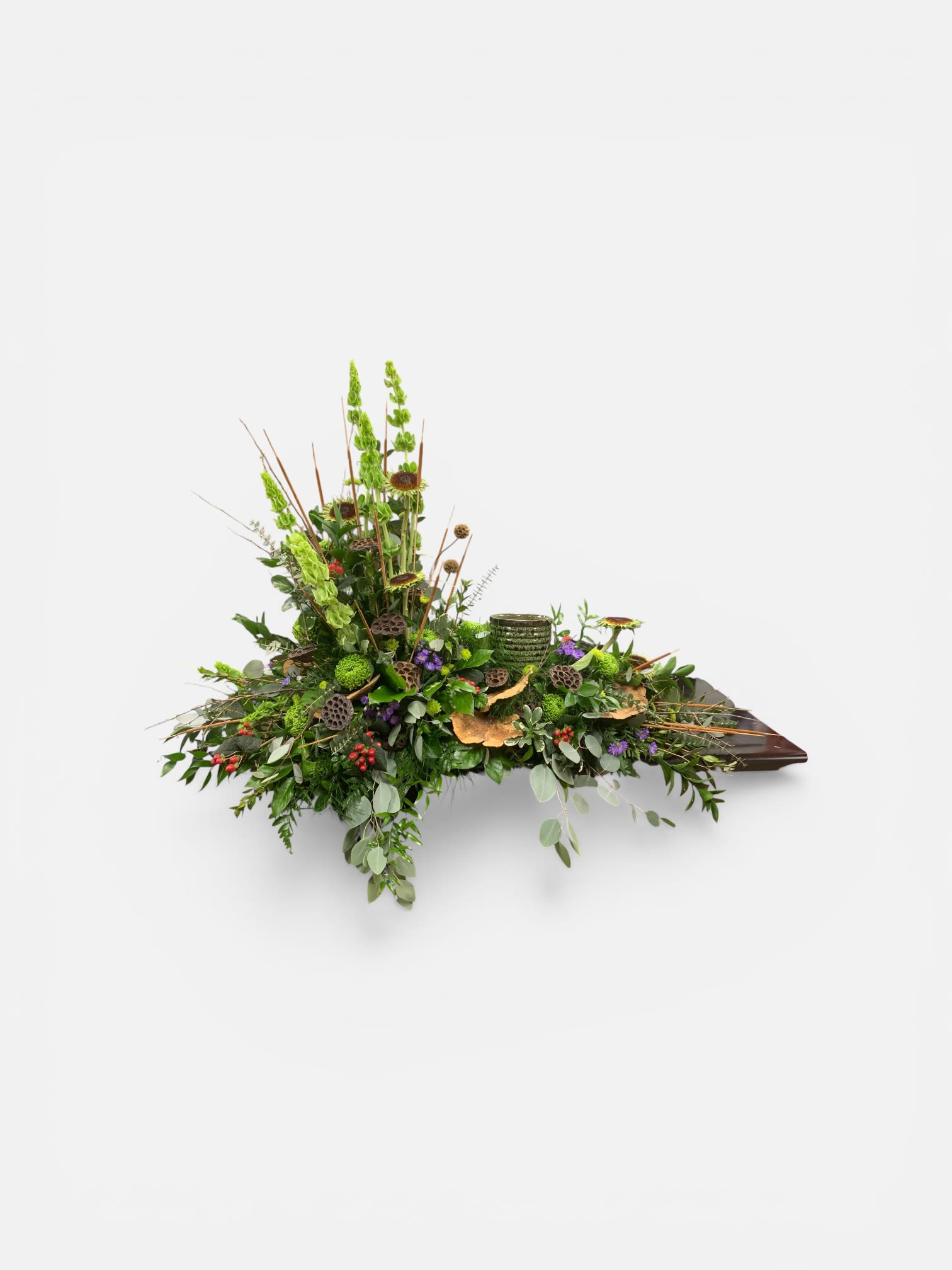 Fields of Green Cremation Wreath  - A graceful, sculptural sympathy arrangement built around tall spikes of Bells of Ireland, this cremation wreath-style floral piece is designed for funerals and quiet remembrances where subtlety matters. The long, low composition spreads horizontally like a field of green, so the person you're honoring will be remembered with a calm, natural statement rather than an overpowering display.  Layers of textured green foliage and button-style chrysanthemums create the lush foundation, while small purple asters and scattered red-mixed accents tuck into pods, berries, and varied leaves for delicate pops of color and interest. The asymmetrical silhouette-height concentrated at one side and tapering along the length-gives a contemporary, organic feel. Everything sits in a dark, boat-shaped ceramic tray that grounds the piece and keeps the arrangement low and dignified for placement on memorial tables or as a centerpiece at a service.  Thoughtful for sympathy, memorials, or a quiet anniversary of loss, this arrangement reads as both respectful and intimate-the kind of piece that helps the recipient feel gently seen. Designed to arrive ready to display, it requires minimal care; keep it out of direct sun and replenish water as needed to preserve the fresh textures and color.