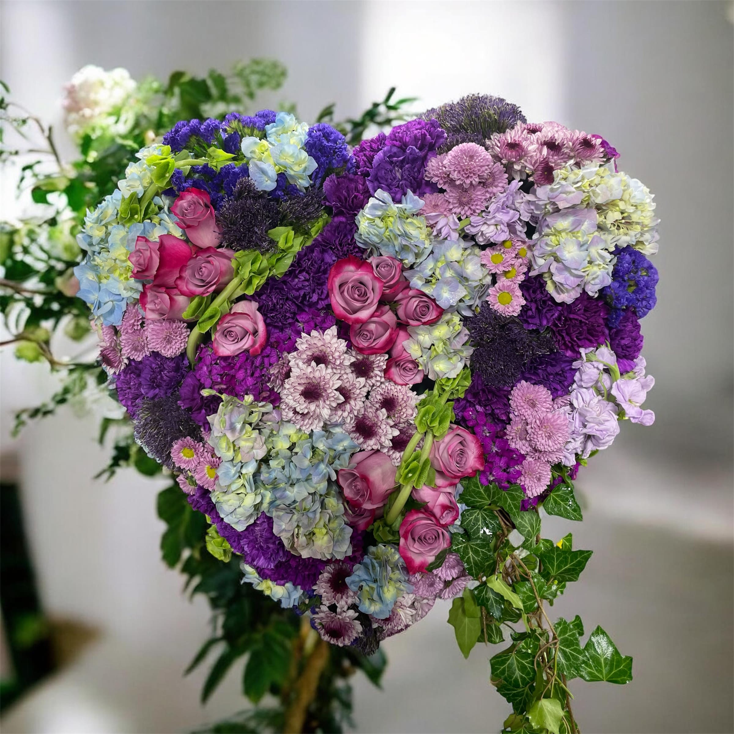 A Love Story  - A plush, heart-shaped floral tribute that speaks of tenderness and memory-the perfect sympathy and funeral arrangement or a deeply sentimental anniversary or Valentine's gesture. This heart-shaped flower arrangement places the recipient at the center, offering a comforting, graceful visual that will make them feel seen and honored in a moment that matters.  Densely packed with lavender, purple, pink and soft-blue blooms, the design features clusters of pink roses nestled among textured cushions of hydrangeas and chrysanthemum petals for rich layers of color and shape. The surface is almost completely covered in flowers for a full, luxurious look, while trailing strands of ivy and green accents frame the lower edges and add a gentle, natural drape.  Ideal for honoring a cherished life or marking a romantic milestone, this arrangement carries a tender, sentimental mood that suits memorial tributes as well as commemorative occasions. Sizes are arranged to maintain the heart form and dense coverage-please contact us if you need specific dimensions or special delivery timing to ensure it arrives looking fresh and arranged for the moment you intend.