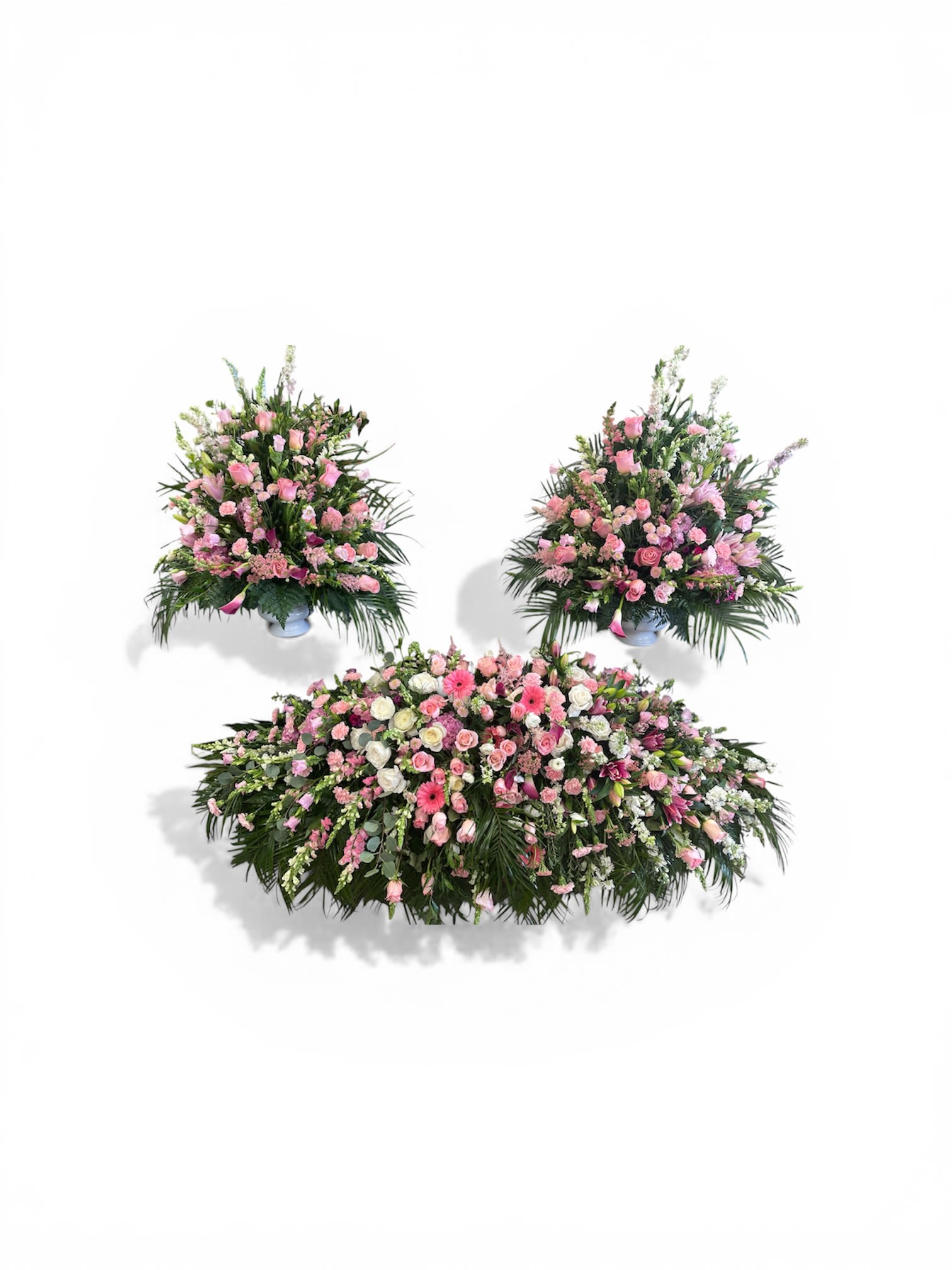 The Sorrowful Goodbye Collection  - Soft pinks, whites, and hints of lavender shape a tender sympathy flower arrangement set designed to offer comfort at a funeral or memorial service. This coordinated three-piece collection includes a long, low casket spray and two matching side arrangements, making it an elegant choice for expressing sympathy or honoring a loved one with a gentle, feminine palette.  The casket spray stretches horizontally with a subtle cascade at each edge and is densely arranged with roses, gerbera daisies, carnations, snapdragons, and chrysanthemums for a lush, layered appearance. Two fan-shaped side pieces rise gracefully from classic white pedestal urns, echoing the same mix of blooms and soft hues to create a unified, formal presentation. The composition balances texture and color-velvety rose petals, bold daisy faces, and ruffled carnations-to read as both reverent and comforting from every angle.  Thoughtfully arranged for easy placement during a service, this set removes the guesswork from planning a meaningful floral tribute. It's a compassionate way to help the person you're honoring feel seen and remembered; sizing and urn details are as shown, and our florists hand-design each piece to preserve freshness and reverence for the occasion.