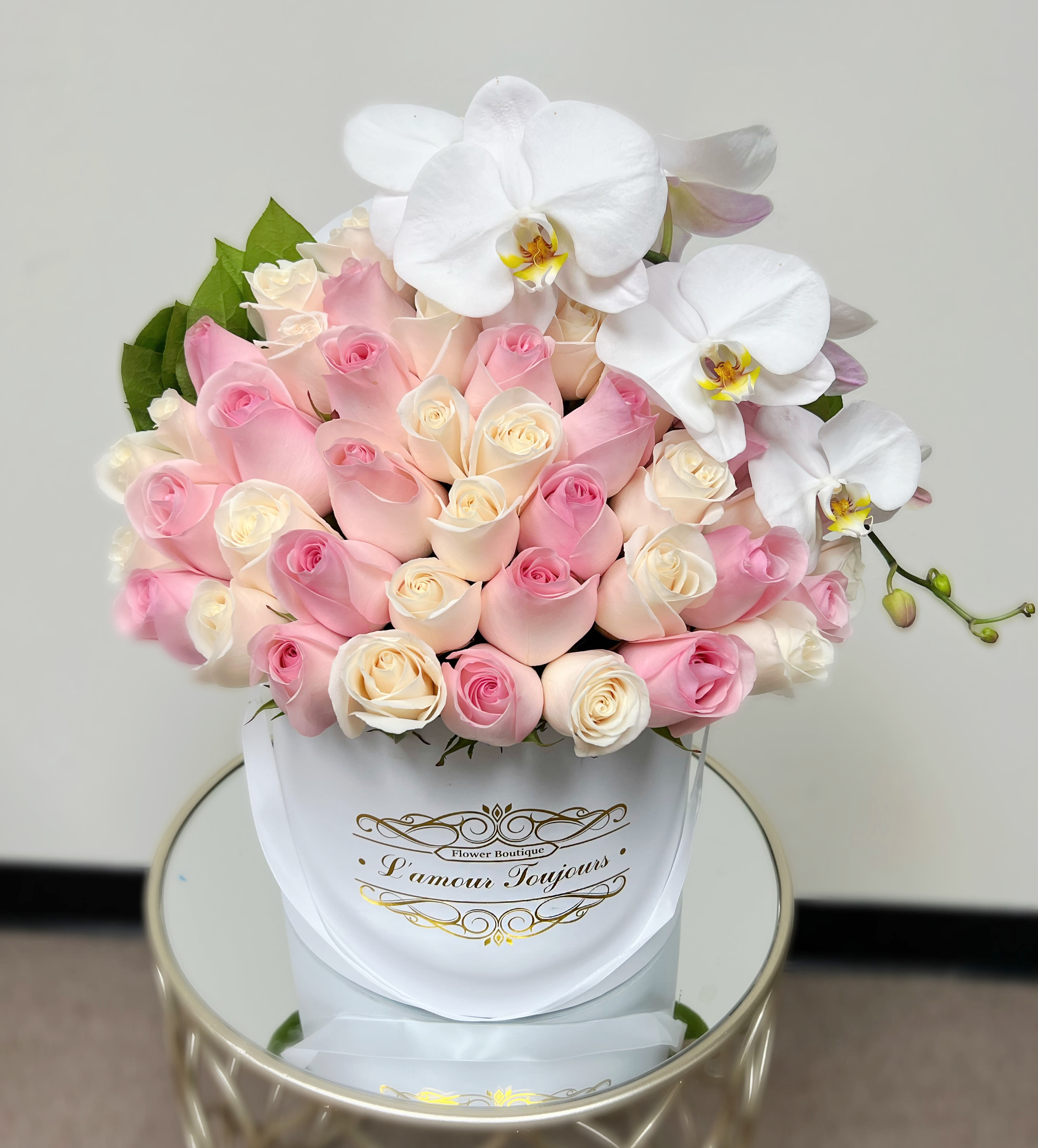  White and Pink Roses with Orchids Signature Box - A lush cluster of blush pink, peach, and creamy white roses forms the heart of this signature box arrangement - a romantic flower arrangement perfect for anniversaries, Mother's Day, birthdays, or simply to say "I'm thinking of you." A sculptural spray of large white Phalaenopsis (cymbidium-style) orchids cascades from one side, creating an elegant contrast in texture that makes the recipient feel truly celebrated.  Hand-presented in a white hatbox with refined gold script, the compact round design sits beautifully on a mirrored side table for a polished, contemporary look that suits weddings, New Baby congratulations, sympathy and funeral settings, or a sophisticated holiday gift. The mix of garden roses, spray roses, and orchids in soft pastels reads as timeless for spring and Easter yet equally lovely through autumn and winter. Ideal for expressing love, gratitude, apology, or get-well wishes, this arrangement is designed to make someone feel seen and cherished.