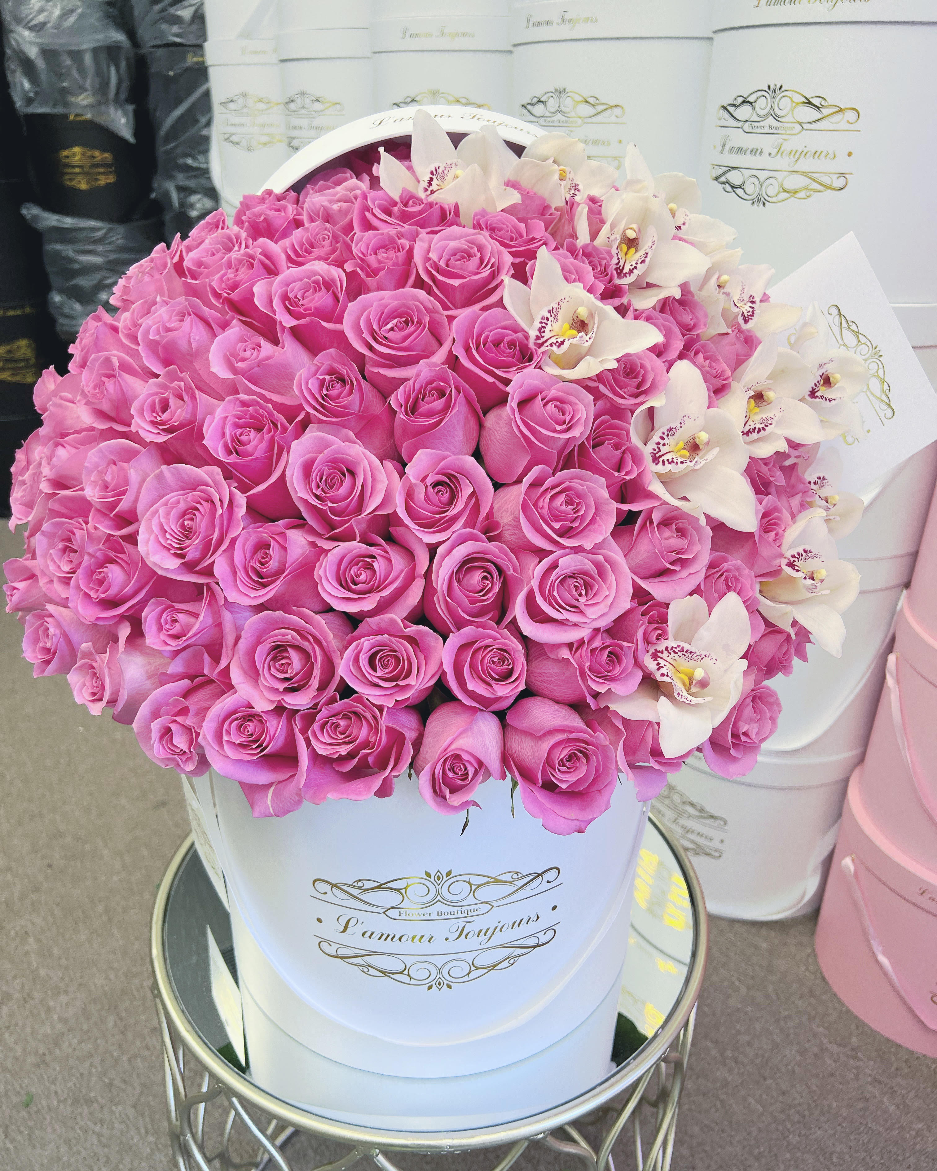 100 Barbie Roses - A lush dome of bright pink roses creates an opulent, romantic statement bouquet perfect for anniversary roses, Mother's Day, or a big birthday surprise. The densely clustered pink blooms read as joyful and celebratory at first glance, while a spray of creamy white cymbidium orchids with speckled centers adds elegant contrast and delicate texture - the kind of arrangement that makes the person you're thinking of feel truly seen.  Arranged in a white round hatbox with gold script and presented on a mirrored metal side table, this composition feels both modern and luxe; a small card is included for your personal message. The compact dome sits low and full, making it ideal for a centerpiece at a wedding, graduation party, or holiday table, and its durable hatbox container keeps stems neatly supported for easy display. Gentle care - fresh water when needed and keeping blooms out of direct heat - will help these roses and orchids stay radiant whether you send them for Valentine's Day, Mother's Day, a get well wish, or simply to say "I'm thinking of you."