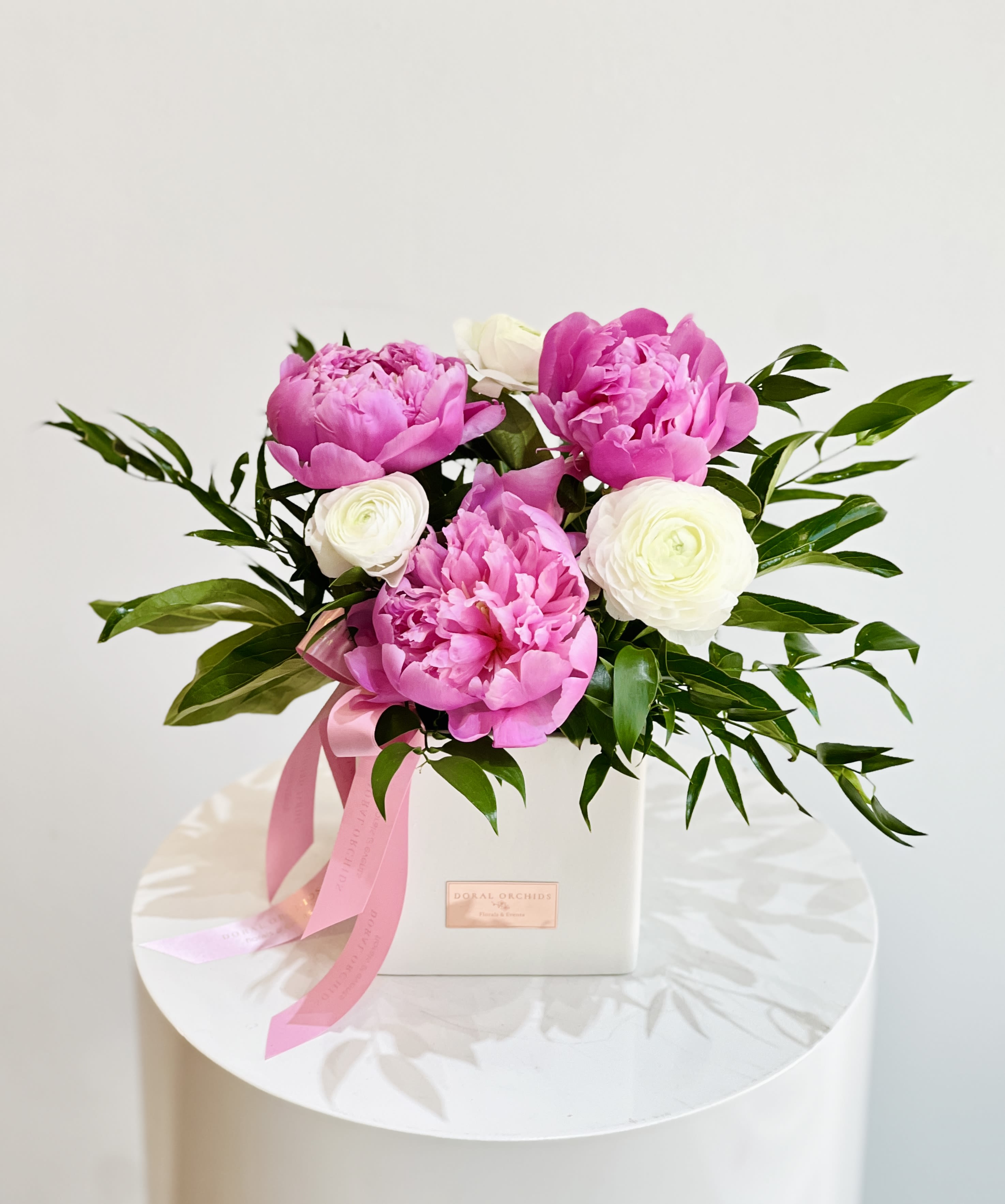 For Her ™ - Elegant arrangement of pink Peonies, white Ranunculus and vibrant Italian Ruscus, in a white ceramic vase.