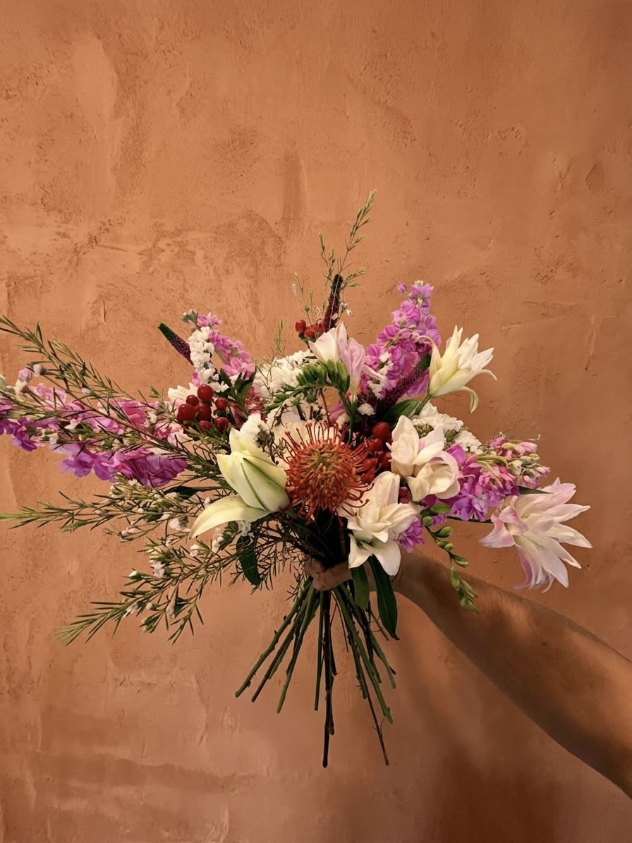 Crown Jewel  - Centered around the architectural brilliance of the Pincushion Protea, The Crown Jewel is a study in botanical contrast and vibrant energy. This sculptural focal point is nested within a lush bed of snowy White Cremones and ethereal Double Oriental Lilies, offering a soft, velvet-like counterpoint to the Protea’s radial form. We’ve layered this arrangement with the sweet, nostalgic fragrance of Freesia and the plush verticality of Stock. Clusters of crimson Hypericum Berries add a playful, beaded texture, while a delicate mist of Wax Flower and Statice provides a fine-grained finish to the silhouette.