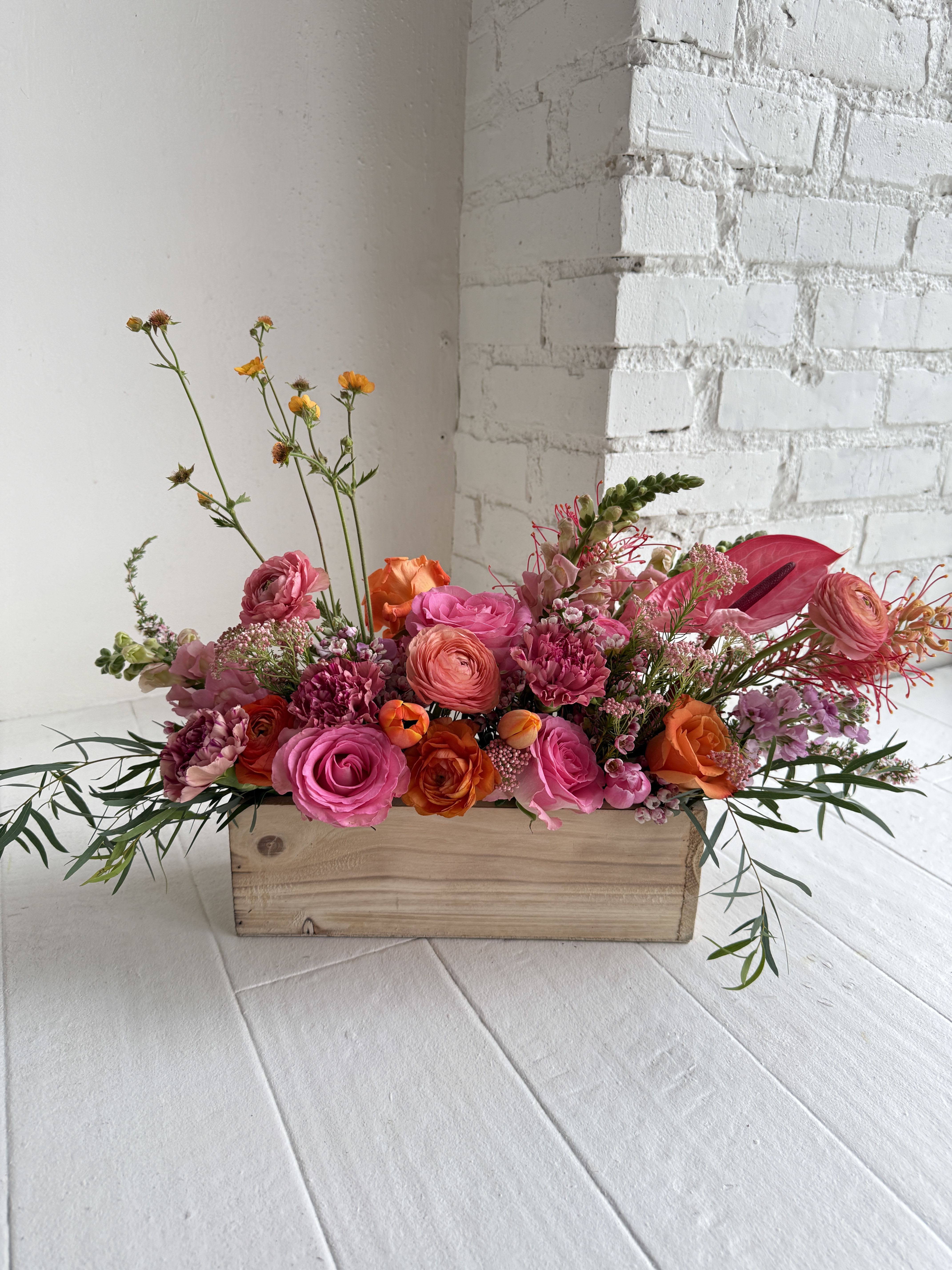 Lola - Bright. Bold. Beautiful. We take our handcrafted wooden crate and all the best blooms to create a lush garden of flowers. Perfect for all sentiments and occasions. This arrangement is low measuring approximately 12" long with an overall height of 8". We favor locally grown in season flowers saturated in shades of pink, peach and orange. Peonies, dahlias, ranunculus, heirloom mums, roses, anthurium, poppies, stock, snapdragon, finished with a fringe of foliage. 