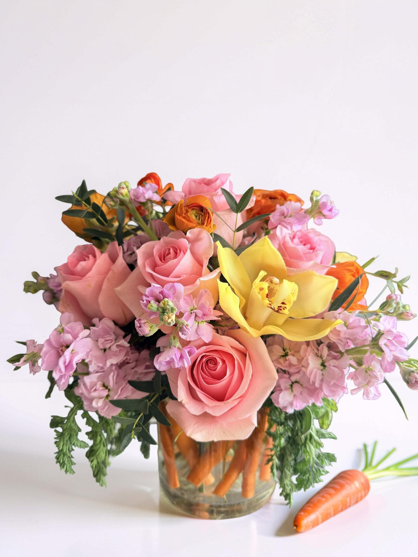 Garden Harvest Glow - A vibrant, garden-inspired arrangement bursting with soft pink roses, cheerful yellow orchids, and delicate blush stock, accented with fresh greenery and fresh carrots . Playfully designed in a clear vase lined with crisp carrots, this piece brings a whimsical farm-to-table charm while still feeling elegant and full of life. Perfect for spring celebrations, brunch tables, or a unique, eye-catching gift.