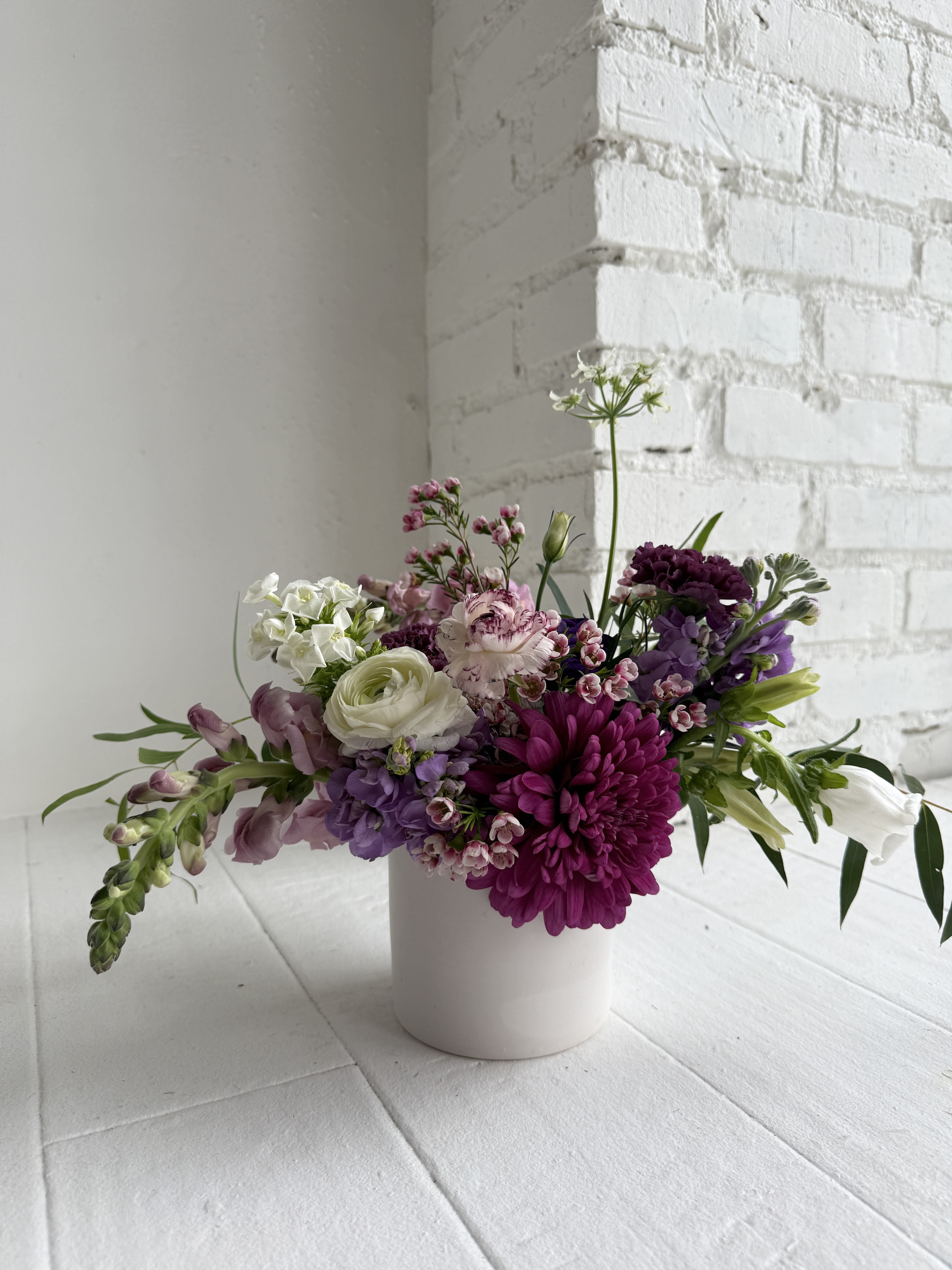 Amethyst - We take a 4x4" white ceramic container and artfully arrange fresh blooms in all shades of violet, purple, pops of white and finished with gorgeous lush foliage. Our flowers reflect the season, favoring ranunculus, roses, stock, snap dragons, anemones, spray roses, wax flower, heirloom mums, carnations, lisianthus, to name a few. This arrangement is perfect for all sorts of occasions including birthday, get well, thank you, baby and just because. The overall size is approximately 10" tall and 6" wide. Upgrades are available for this arrangement at check - out. We promise your arrangement will be stunning, fresh, unique and thoughtfully crafted using as many locally grown flowers as possible.