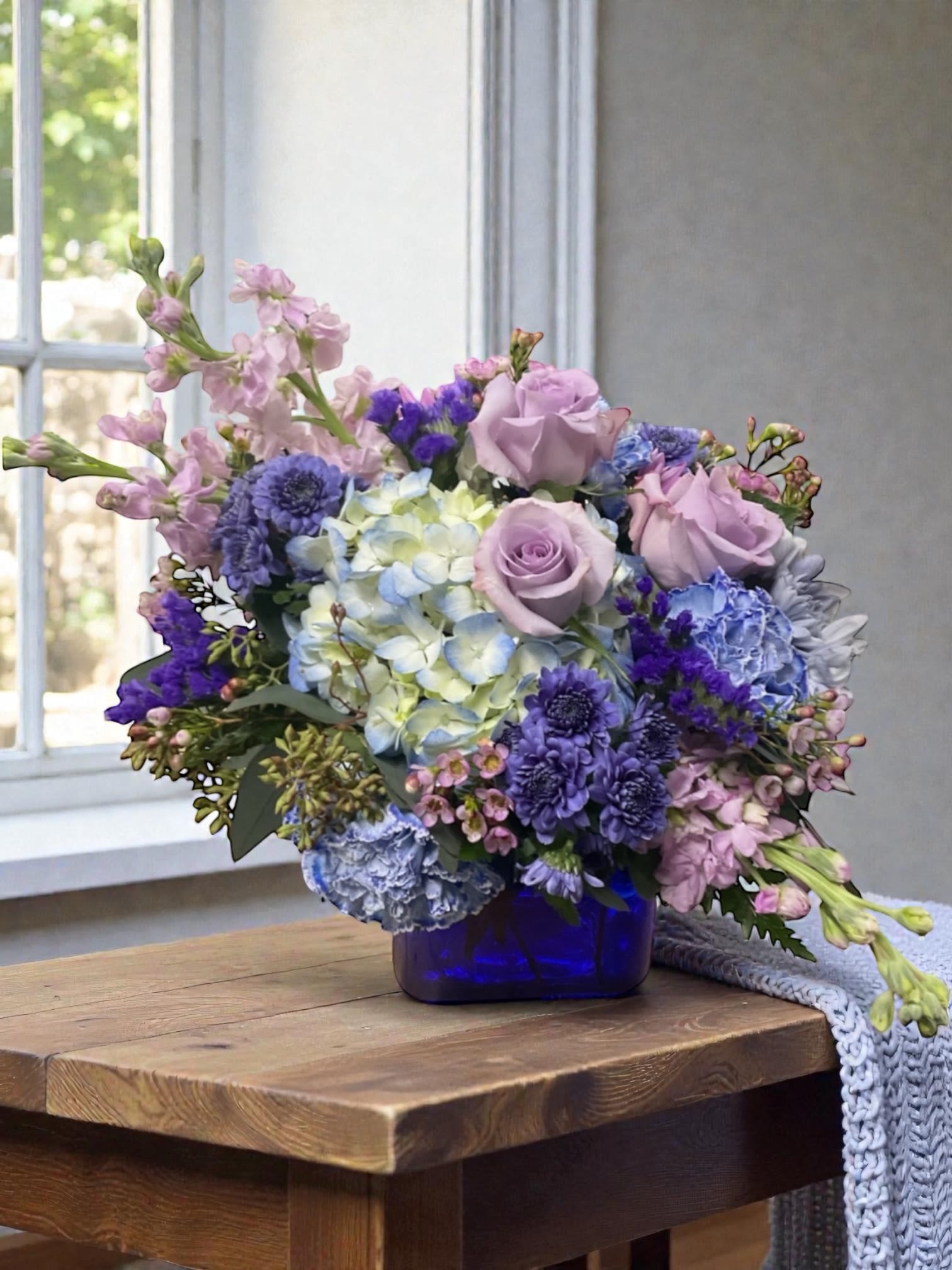 Midnight Garden Arrangement  - Midnight Garden is a modern statement arrangement designed in a bold cobalt blue cube, featuring lavender roses and lush blue hydrangea. The cool, saturated palette creates immediate visual impact while still feeling polished and composed.  The compact cube style keeps the design structured and intentional, allowing the color to take center stage without feeling busy or overworked. It’s a look that feels confident, elevated, and distinctly modern.  A standout choice for birthdays, thank-you gifts, thinking of you, or everyday moments when you want to send something with a little more presence.  Each arrangement is designed with the freshest stems available. Varieties may vary slightly while maintaining the same color palette and overall style.  Available for local flower delivery through Aspen Tower Florist, serving Shelbyville and Greensburg along with Rushville, Batesville, Westport, Oldenburg, St. Paul, Waldron, Milroy, and Hope.