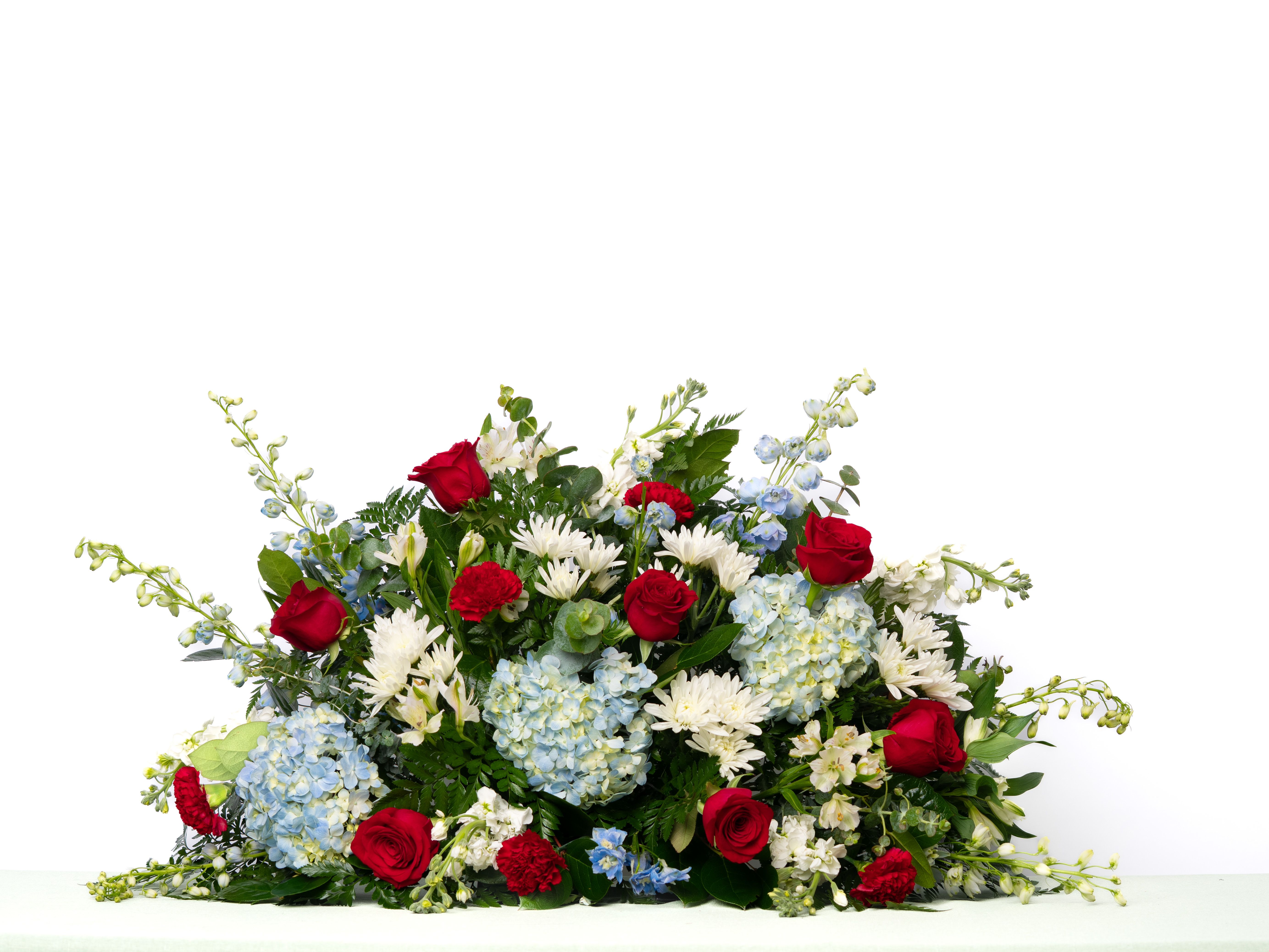 Our Honor Collection: Half Casket Spray in Patriotic Red, White, and Blue Blooms - A patriotic tribute honoring dedication, service, and legacy. Designed in bold red, white, and blue blooms, this arrangement offers a respectful and meaningful way to commemorate a loved one.  Each arrangement is thoughtfully designed using the freshest blooms available. Flower varieties may vary based on availability while maintaining the overall style, color palette, and value.  Approximate Dimensions of Floral Tribute: 20" tall x 36" wide  Color &amp; Customization: Need a custom color scheme? Contact us for more information. Our most popular palettes include whites, whites and greens, whites and blues, pinks and whites, pastel shades, and colorful designs.  Delivery: All sympathy pieces are hand-delivered by the Schultz Floral team.