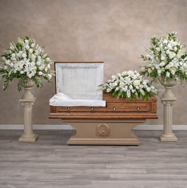 Eternal Grace Set - A serene all-white sympathy arrangement designed to honor a life with quiet dignity - perfect for a funeral or memorial service. The Eternal Grace Set features a low, elongated casket spray composed of white lilies, roses, carnations, chrysanthemums, stock, and snapdragons that forms a soft horizontal line across a wooden casket, while matching companion pieces lend a formal, balanced presence.  Two large companion arrangements in urn-style vases sit on tall pedestal stands at either side of the casket, with simple greenery to accent the white blooms and keep the focus on their pure tones and gentle textures. The coordinated set creates a reverent, peaceful atmosphere that helps the person you're thinking of feel cherished and remembered; sizing and stand arrangements are designed to fit standard casket displays for a cohesive presentation.