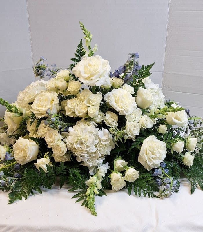 Peaceful White Casket  Spray - A serene cascade of white roses, spray roses, and full hydrangeas forms the centerpiece of this white casket spray, a sympathy arrangement designed to convey quiet comfort and respect. The low, elongated mound and gently rounded top create a formal, structured silhouette ideal for memorial services and funerals, while soft blue delphinium stems tucked through the composition lend a cool, peaceful contrast that feels calming rather than showy.  Carefully layered greenery frames the edges, giving the piece a dignified border and subtle texture that complements the trailing white snapdragons and upright snapdragon accents. Chrysanthemums add fullness and longevity to the bouquet, so the flowers maintain their presence throughout the service. The overall palette of white, blue, and green keeps the tribute understated and elegant.  Made to lay gracefully atop a casket or to serve as a focal sympathy tribute, this arrangement offers a meaningful, composed way to honor someone's memory. Sizes are crafted to standard casket dimensions-please consult our staff for specific measurements or custom requests-and we recommend keeping the spray cool and out of direct sunlight until presentation to preserve freshness.