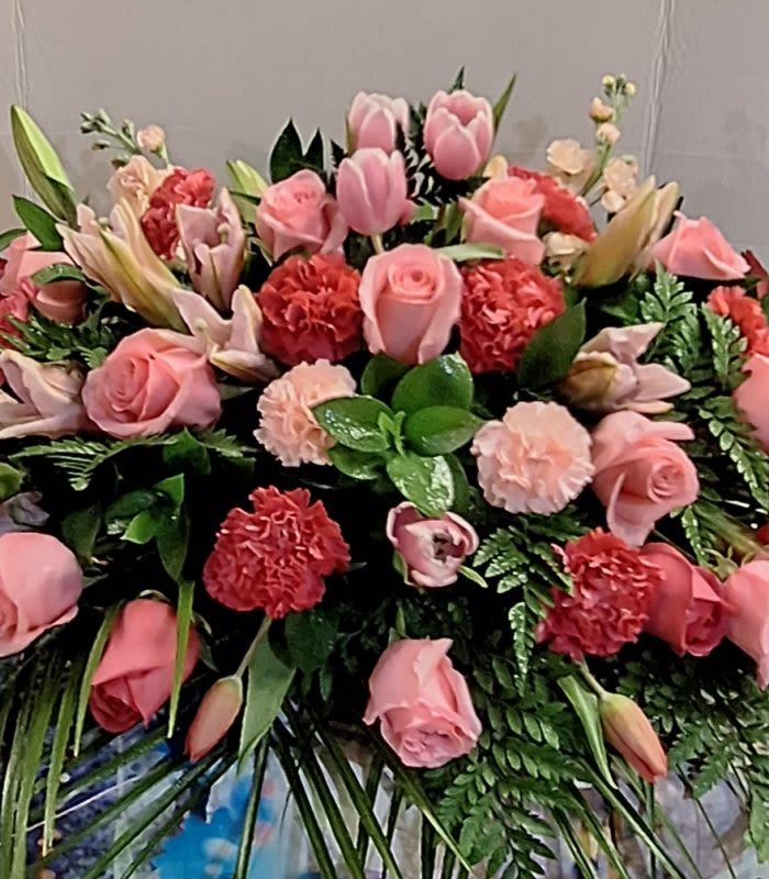 Remembering Her Casket Spray - A lush, rounded spray of pink and red blooms arranged over glossy greenery offers a tender, heartfelt tribute for sympathy and funeral services. This casket spray blends soft pink roses and tulips with pale lilies and ruffled carnations-accented by deeper red carnations-to create a warm, romantic palette that reads as both comforting and celebratory of a life remembered.  The design is densely massed, fanning outward from a base of assorted greenery and long palm-like fronds for a full, sculpted silhouette. Colors range from pale peach-pink to vivid red, and textures move from the smooth petals of roses and tulips to the frilled edges of carnations and the elegant trumpet shape of lilies, producing a rich, layered look appropriate for placement on a casket or as a prominent display piece.  Arranged to convey affection and care, this spray is a meaningful way to honor a loved one and offer solace to family and friends. Available in a full size to suit most casket presentations; please contact us for delivery timing and any personalization requests to ensure on-time placement for the service.
