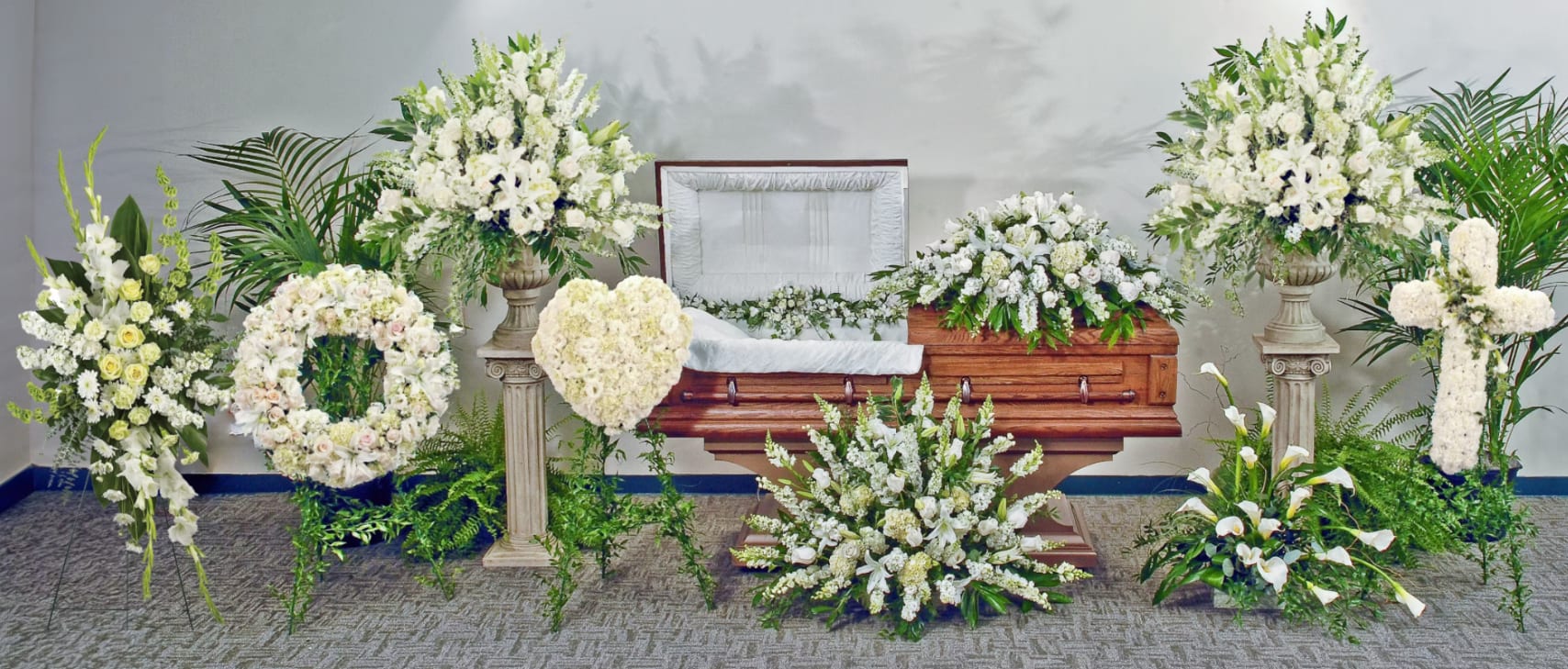 Eternal Grace Collection - A serene all-white funeral arrangement designed to honor a loved one with quiet dignity-this sympathy floral ensemble is ideal for a funeral or memorial service. The display centers on a wooden casket draped with a large casket spray surrounded by coordinating floor sprays, so the person you're remembering is framed by soft white roses, lilies, calla lilies, chrysanthemums and complementary blooms that convey respect and calm.  Tall, overflowing arrangements on stone pedestal stands anchor each side of the casket, while rounded wreath and heart pieces rest on metal easels to offer traditional symbols of remembrance. A cross-shaped design and groupings of white calla lilies extend the theme, and lush green foliage and potted plants form a rich backdrop that highlights the purity of the white flowers and gives depth to the presentation. The mix of textures-from the sculpted roses to the graceful calla lilies-creates a balanced, reverent look appropriate for both intimate services and larger memorials.  If you're arranging a service, this coordinated grouping sends a clear message of sympathy and honor to family and friends and helps focus the ceremony on remembrance. Available as a full-service presentation with pedestal stands, easels, and casket spray already matched for color and style, these pieces are prepared to set up at the venue for a respectful, cohesive tribute.
