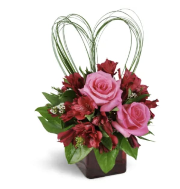 Blush of Devotion - Sweet, stylish, and full of romance, this arrangement features soft pink roses nestled among rich red blooms and fresh greenery. Designed in a modern cube vase with a delicate heart accent above, it’s a charming expression of love—perfect for making someone feel truly special.
