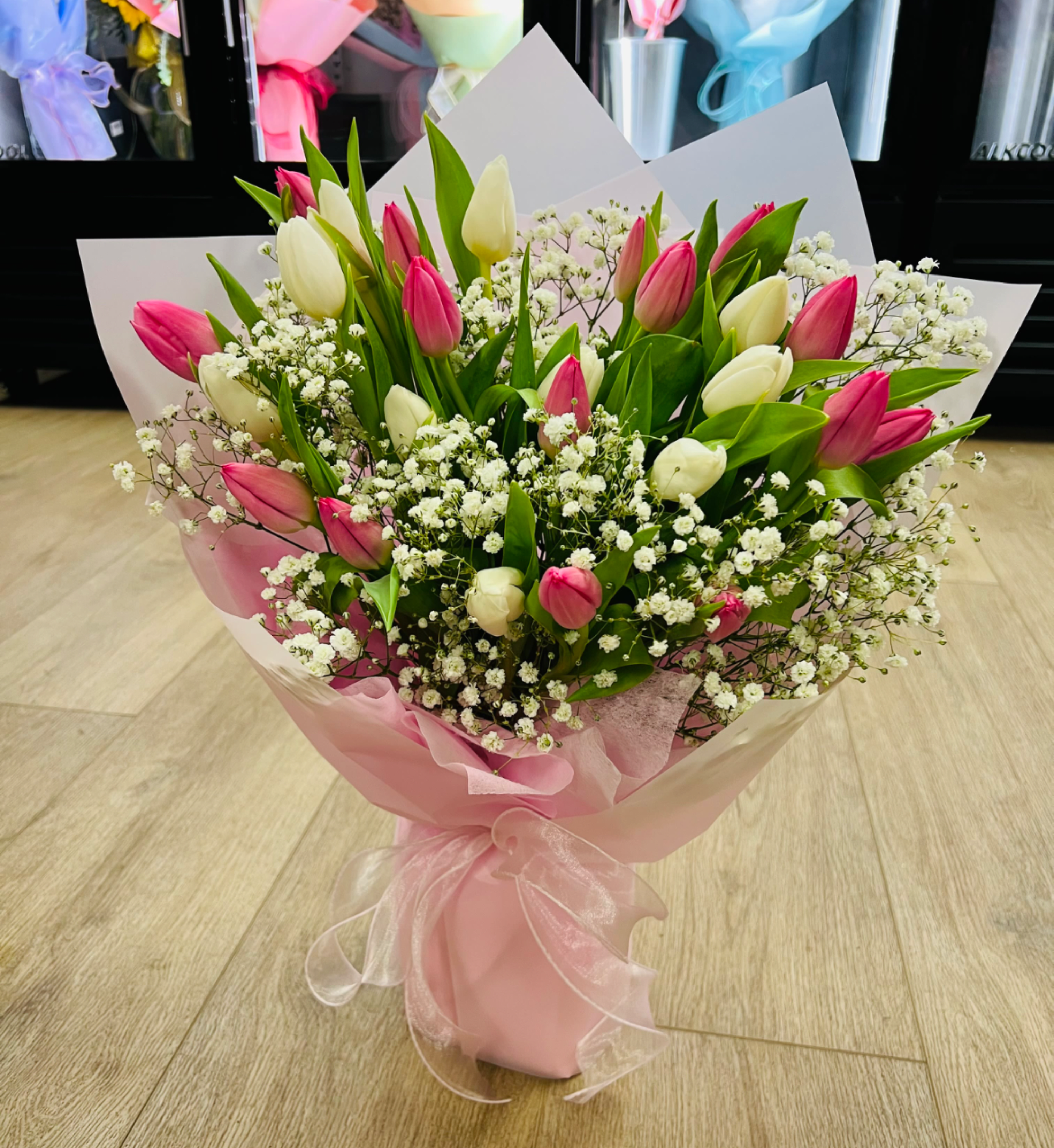 Hand-tied bouquet of pink and white tulips with small white filler flowers wrapped in pink paper and ribbon.