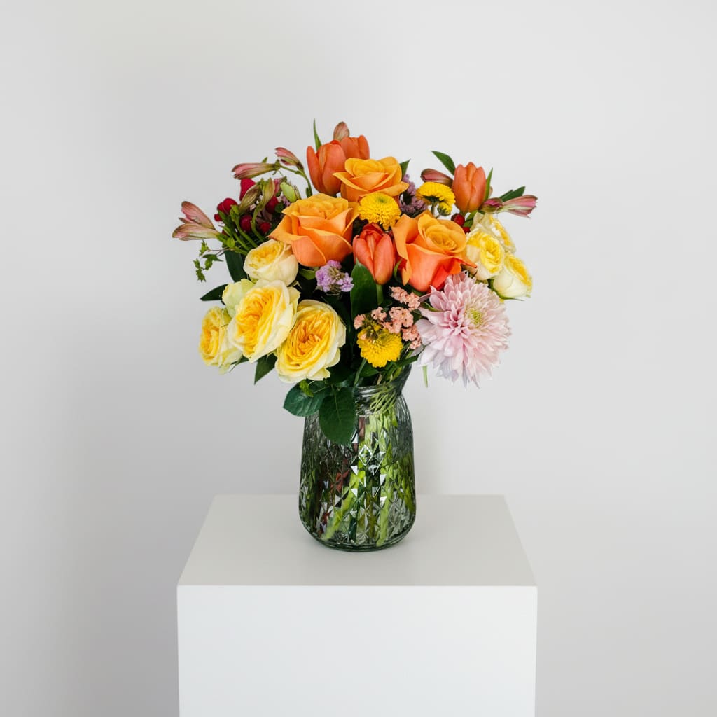 Joyful  - Sunny orange and yellow roses paired with bright orange tulips make an immediate, radiant impression in this cheerful flower arrangement for get well wishes, thank-you gestures, or a just-because surprise. The warm autumnal palette-accented by soft pink dahlias and blush alstroemeria-reads as both celebratory and comforting, the kind of bouquet that will make the person you're thinking of feel truly seen and uplifted.  A lively mix of roses, tulips, chrysanthemums (button mums), dahlias, alstroemeria, and stock is arranged in a textured green patterned glass vase (also offered in a clear patterned glass option), letting stems peek through for a classic, homey look. The rounded, dense grouping balances bold yellow and orange with pastel fillers and fresh greenery for pleasing texture and scent; dimensions are designed to sit happily on a bedside table, desk, or entry console.  Perfect for autumn celebrations like Sukkot but equally appropriate year-round, this arrangement is ideal when you want to send warmth, encouragement, or thanks. Handcrafted from fresh, high-quality blooms, it arrives ready to display-just add water and enjoy the bright, long-lasting color.