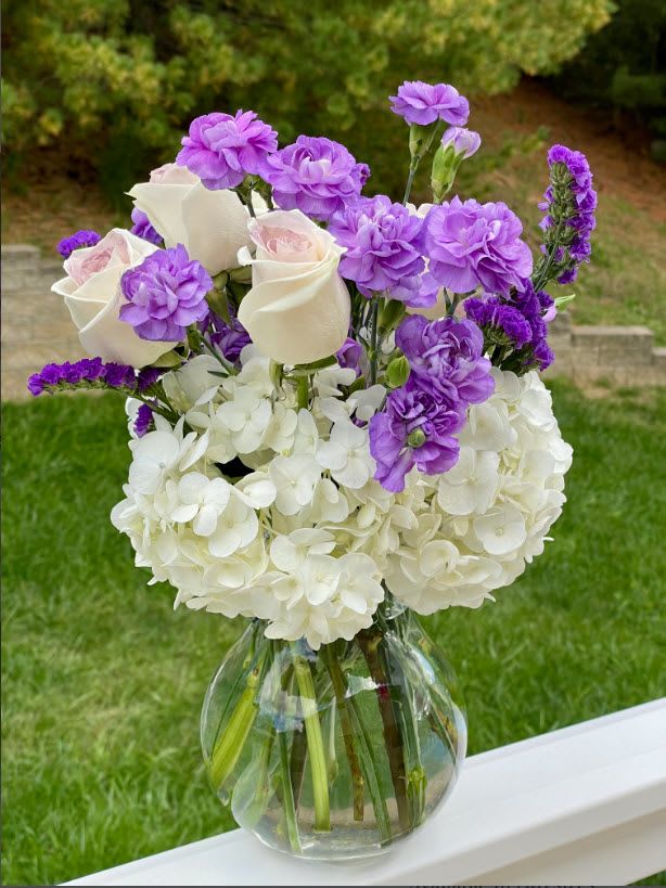Lavender Elegance - This elegant bouquet features a soft blend of white hydrangeas, creamy roses, and vibrant purple carnations, accented with rich purple statice flowers. It's designed to add a touch of sophistication to any space or occasion.   Hydrangeas Carnations Roses  Original: Includes six creamy roses, offering a charming and thoughtful gift option while still capturing the bouquet's beauty.  Arranged in a classic round glass vase, this bouquet is a timeless centerpiece or gift for any celebration.  Height 11 inch Width 9 inch