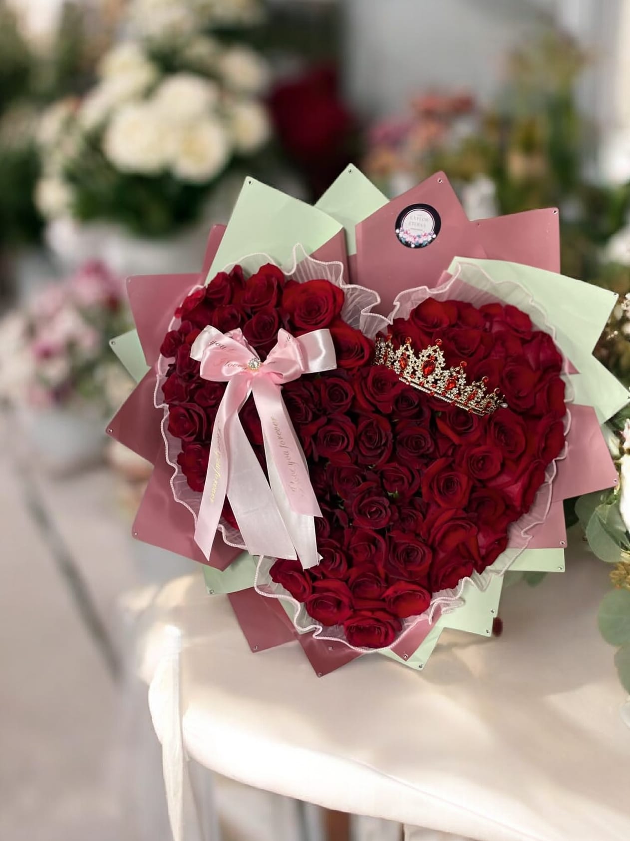Corazon Mio - A dense heart of deep red roses shaped into a classic heart makes an unmistakable romantic statement-perfect as a birthday bouquet, anniversary roses, or a heartfelt tribute for someone you adore. The arrangement reads like a keepsake gift: her reaction will be the focal point, whether you're celebrating love, showing appreciation, or marking a special milestone.  The roses sit in a heart-shaped box and are edged with soft white ruffled trim that highlights the heart silhouette against layered pastel pink and mint panels. A satin pink ribbon bow and the roses for a celebratory,; decorative wrapping paper completes the polished presentation. Sized and styled to be displayed as a centerpiece or special keepsake, this arrangement arrives ready to present and makes the recipient feel truly celebrated.
