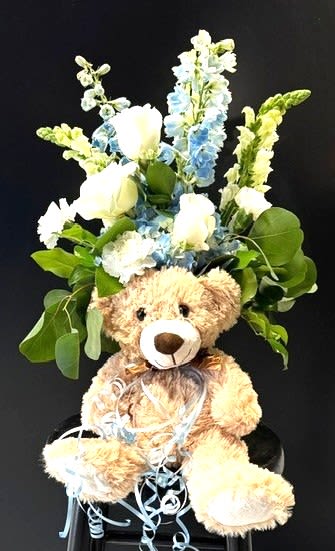 #A009 - Snips, Snails, &amp; Puppy Dog Tails - Little boys are such a joy and our Snips, Snails, &amp; Puppy Dog Tails celebrates what makes them tick!  A cuddly tan teddy bear tucked before a tall, sky toned floral spray creates a playful, welcoming centerpiece perfect for a new baby or baby's first birthday. This vertical birthday arrangement and new baby gift pairs soft blue delphinium spikes and pale snapdragons with clusters of white roses and carnations to give the recipient a sweet, celebratory surprise that feels both joyful and comforting.   These colorful blooms and generous green foliage form a garden like backdrop that frames the bear and adds airy texture, while light blue curling ribbon at the base brings a whimsical finishing touch. Approximately life sized for a nursery table or hospital bedside, the mix of long stems and rounded blossoms gives good visual height and keeps the plush companion front and center - the kind of gift that will make a new parent or little one feel truly special.