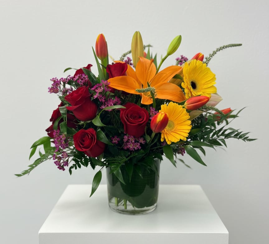 Colorful arrangement of red roses, orange lilies, and yellow gerbera daisies in a clear glass vase