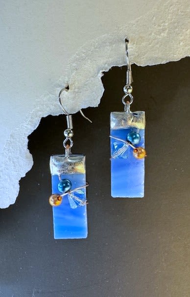 #A024 - Sky Blue Stained Glass Earrings - A pair of sky-blue stained glass dangle earrings that catch the light with soft horizontal bands of blue-perfect as a birthday gift, Mother's Day surprise, or a thoughtful "just because" for someone who loves handmade accessories. These rectangular glass drops feel modern and artistic while remaining lightweight for everyday wear; they read as both playful and polished for occasions from anniversaries and graduations to a sweet holiday stocking stuffer.  Each earring is finished with a silvery metal cap and metal earring hooks, accented at the front with small round beads in teal and amber plus clear crystal-like pieces wired in for extra sparkle and texture. The translucent glass shifts from pale to deeper blue across the piece so the color changes with movement and light; overall length and exact dimensions are ideal for comfortable dangle without weighing the ear. Handmade in our shop in the Shenandoah Valley, these make a lovely add-on to a bouquet, a small standalone gift for prom or wedding parties, or a "get well" or "thinking of you" token that feels personal and well-made.