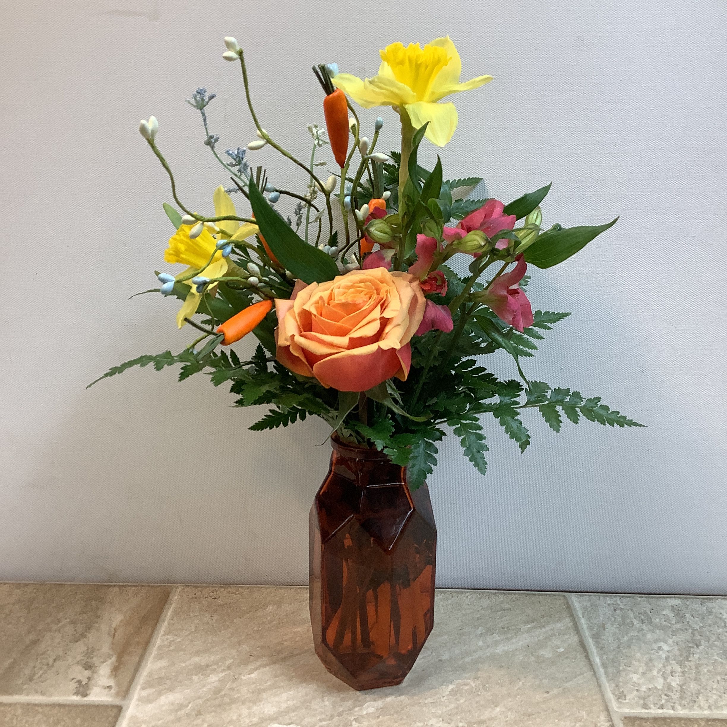 Carrot Patch - This bouquet has a bright, cheerful springtime feel with a playful twist. At the center sits a warm peach rose, its soft, glowing petals creating a gentle focal point. Surrounding it are sunny yellow daffodils that add a burst of light and movement, paired with delicate pink alstroemeria for a touch of sweetness. Whimsical accents of small decorative carrots and airy filler flowers weave throughout, giving the arrangement a fresh-from-the-garden charm. Lush greenery and fern fronds provide a rich, natural base, all gathered in a warm amber glass vase that enhances the cozy, rustic look. It measures 10” wide x 15” tall.