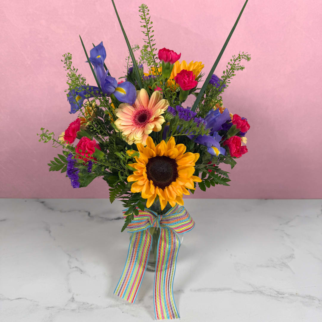 Bright Spring Morning - Bright Spring Morning by Savilles Florist is a cheerful flower arrangement perfect for birthdays, congratulations, or a get-well lift. It's the kind of bouquet that makes the person you're sending it to feel genuinely celebrated. Bold sunflowers take center stage while vivid purple irises, hot pink carnations, and soft peach-pink gerbera daisies surround them, creating an energetic, eye-catching mix that reads joyful from across the room.  The stems are gathered in a clear glass vase and finished with a wide multicolored striped ribbon tied in a playful bow, echoing the bouquet's sunny yellow, deep purple, pink and blue accents. Smaller filler blooms and airy greenery add texture and height so the arrangement looks full and lively from every angle. This is the ideal arrangement for brightening someone's day or celebrating a happy moment in someone's life.