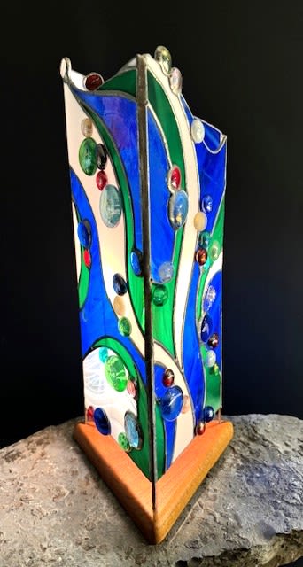 #A044 - Abstract Lamp - A vertical stained-glass sculpture that reads like a ripple of sea glass-this stained glass lamp makes a memorable gift for Mother's Day, a housewarming, or an anniversary. The abstract lamp and art panel style piece functions as both decorative accent and mood light, so the person you're thinking of will feel seen by something both beautiful and distinctly personal rather than a typical bouquet. Handmade three-sided stained-glass panels rise from a triangular wooden base, where flowing bands of blue, green and soft white glass are separated by metal lines for a crisp, modern silhouette.   Round glass gems in mixed colors are scattered across the surfaces, adding jewel-like highlights and tactile dimension; when lit the glass gives a warm, artistic glow that changes with the room's light. The compact vertical form fits easily on an entry table, office desk, or living-room shelf as a sculptural focal. Perfect for birthdays, Father's Day, graduation, retirement, thank-you or "just because" moments, this piece works year-round-from spring refreshes to cozy autumn and winter accents-offering lasting presence instead of fleeting blooms. Choose it when you want a lasting keepsake that brings color and light to a special someone's home; simple care is limited to dusting and positioning where its glow can be enjoyed.