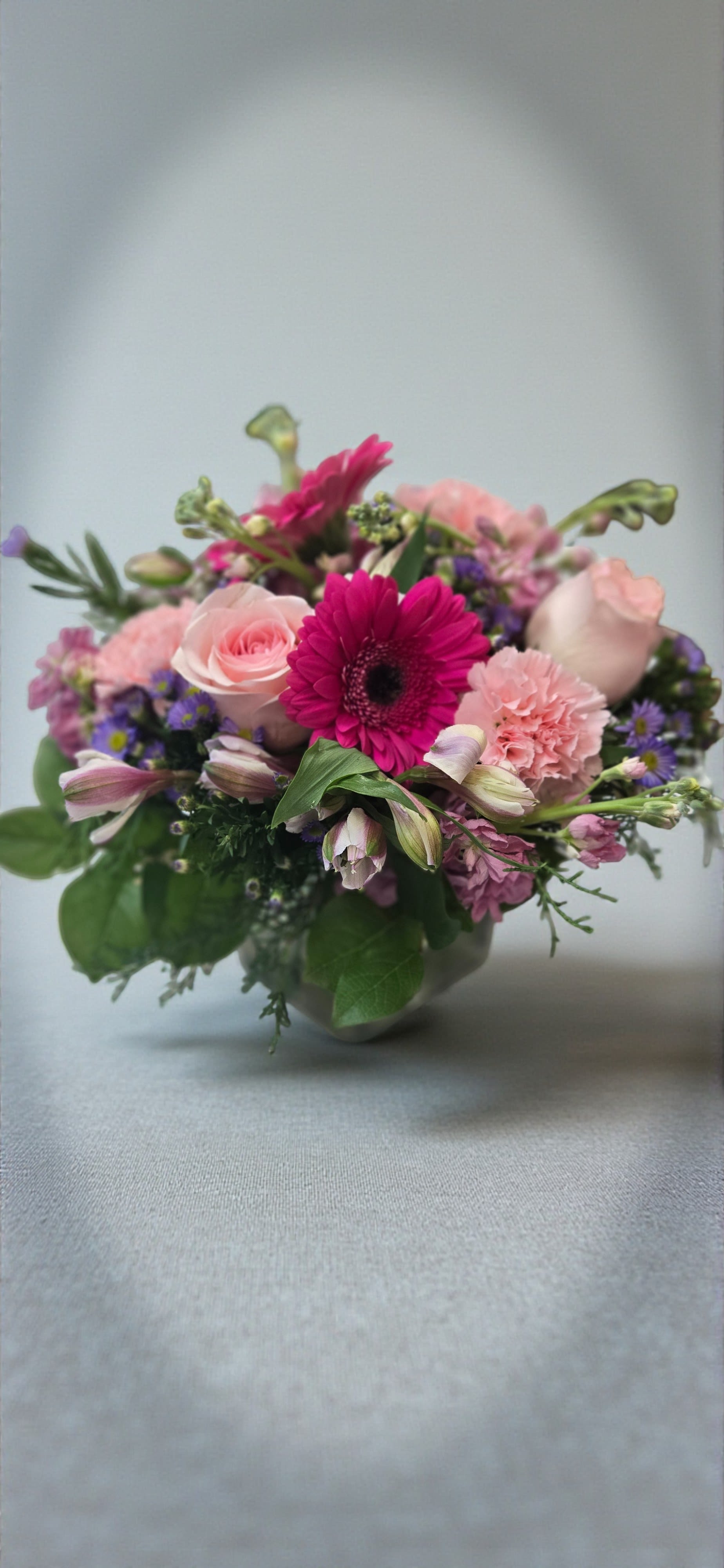 Art &amp; Flowers Exclusive "Loves Garden" - Bright magenta gerbera daisies and blush roses create an immediate "wow" - a garden-fresh anniversary bouquet that also reads perfectly for birthdays, just-because surprises, or a romantic thank-you. This compact flower arrangement is designed to make the person you're thinking of feel seen: the vivid pinks and soft peaches offer warmth, while pops of lavender and white keep the mood light and joyful.  Hand-gathered and low in profile, the bouquet mixes gerbera daisies, roses, spray roses, carnations, alstroemeria, stock, snapdragons, chrysanthemums, hydrangea touches and delicate aster and filler blooms for a layered, textural look. One version is arranged in a clear glass cube vase lined with a decorative green wrap that gives the base a modern, saturated color block; another comes in a clear glass bowl vase with a dense, rounded composition. Fresh green foliage adds airy balance and natural movement across the design.  Ideal for Spring gifting, Women's Day, Best Friends Day, Admin Professionals' Week, or Grandparents and Parents Day, this arrangement is the kind of bouquet that brightens a hospital room, centerpieces a dining table, or says "congratulations" without words. The compact size makes it easy to place and enjoy; simply change the water every few days and trim stems for the longest-lasting vase life.