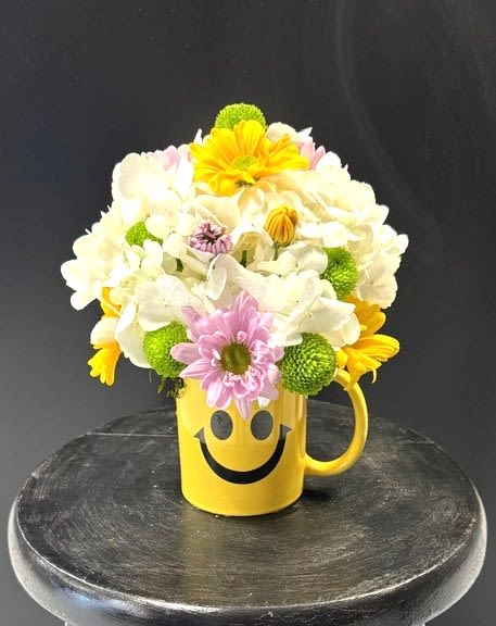 #A054 - Sunshine To Go! - A bright dome of white hydrangeas crowned with yellow, lavender and green daisy-style chrysanthemums makes this flower arrangement an instant mood-lifter - perfect as a Mother's Day bouquet or a cheerful birthday, get well, or a “just-because” surprise. The smiley mug and cheerful flowers fit naturally with most occasions like anniversary, graduation, housewarming, and “thinking of you” – giving your gift a sunny, optimistic message the recipient will notice first.   Flowers are gathered tightly into a compact, rounded shape that sits low and full over the mug, so the bold white hydrangea mass reads like a soft cloud punctuated by pops of yellow, pink-toned lavender and fresh green mums. Chrysanthemums, daisies and hydrangeas combine for long-lasting texture and color contrast, while the yellow ceramic mug with its black smiley face adds a reusable keepsake element that feels casual and joyful. This arrangement is a friendly way to say thank you, congrats, I'm sorry, or welcome a new baby - and it works equally well for Father's Day, retirement, summer celebrations, or sending warm wishes to someone recovering. The compact size makes it table-ready and easy to display; refresh water every other day and trim stems as needed to extend freshness. Order when you want the person you're thinking of to feel genuinely seen and brightened.