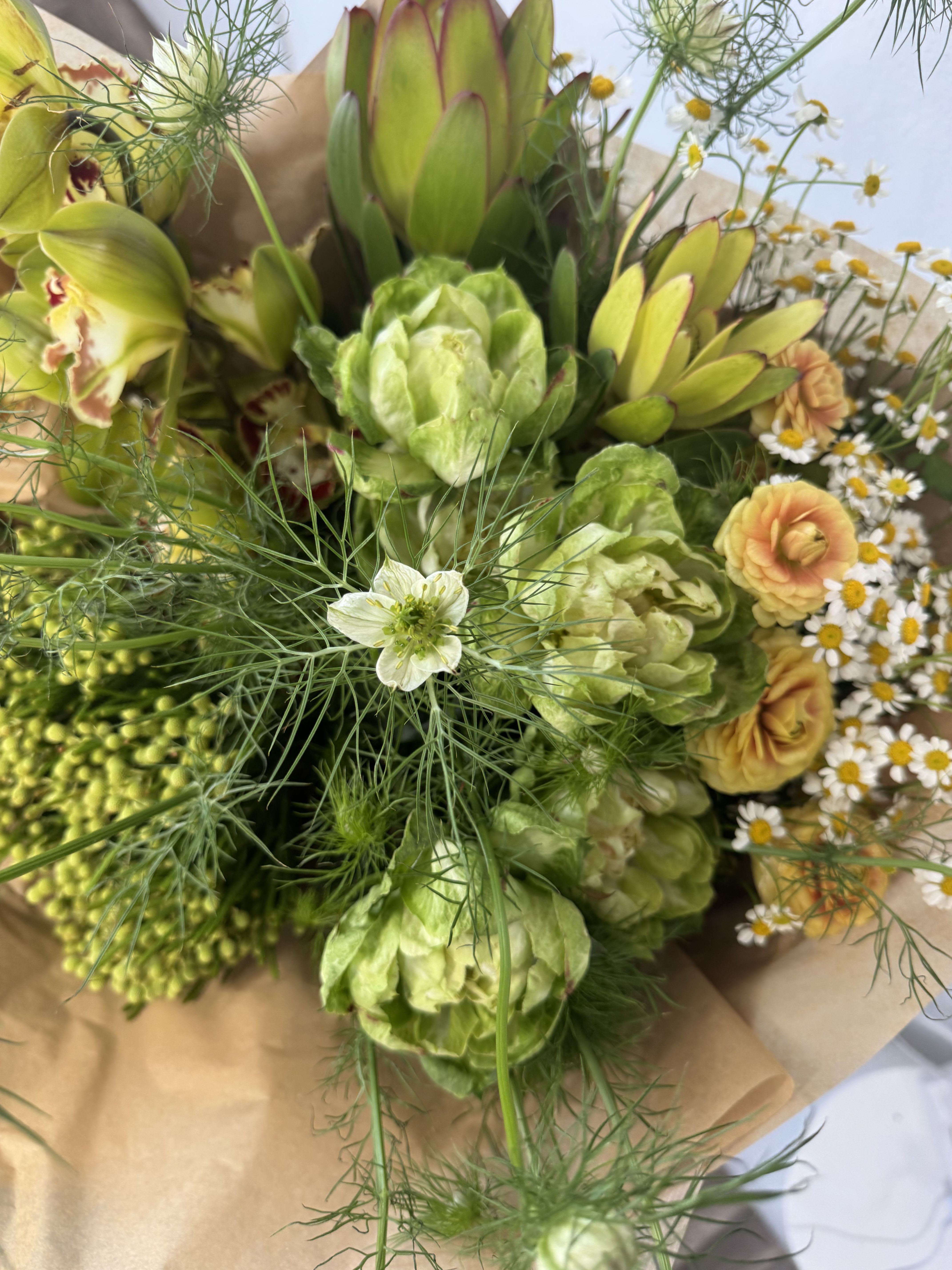 Garden to Table - Bright, garden-fresh design: a hand-tied flower arrangement perfect as a cheerful birthday bouquet or a "just because" gift that feels freshly picked from the greenhouse. The mix of yellow ranunculus and small white daisy-like blooms creates an uplifting, sunny impression, while green protea-style heads and cymbidium orchid blossoms with burgundy centers add unexpected texture and an exotic touch. This arrangement reads as both celebratory and relaxed-the kind of bouquet that makes the person you're thinking of feel noticed and genuinely delighted.