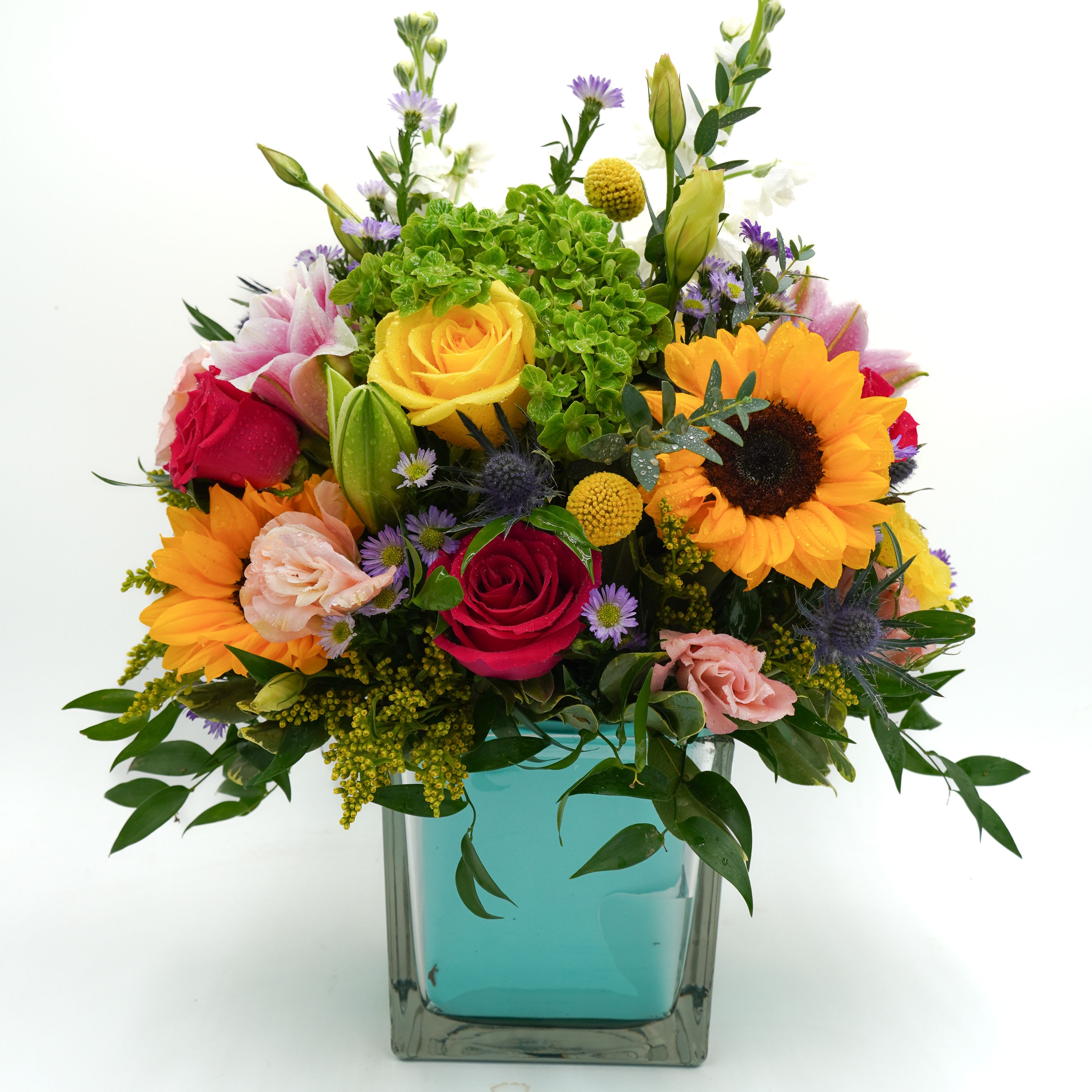 Golden Garden Glow - Brighten any space with this vibrant, garden-inspired arrangement featuring radiant sunflowers, classic roses, soft lilies, and fresh seasonal blooms. Designed in a modern glass vase, its mix of warm yellows, rich reds, and soft pastels creates a cheerful and elegant look—perfect for celebrations, thank-you gifts, or simply bringing sunshine indoors. 