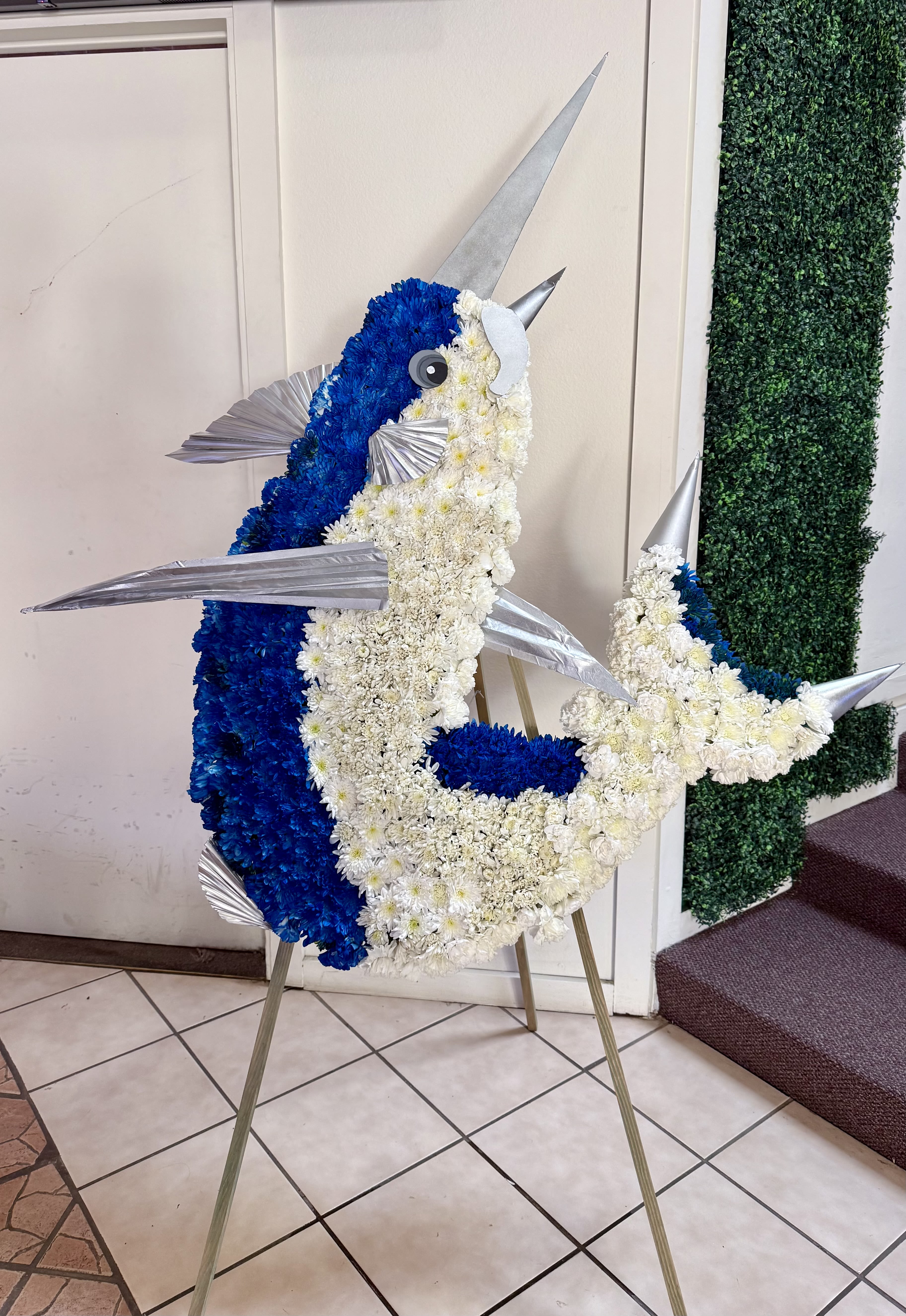 Ocean of Memories - A striking and artistic ocean-inspired floral tribute, designed to honor a loved one with creativity and meaning. This unique arrangement features a beautifully crafted fish silhouette, brought to life with fresh white and deep blue flowers, symbolizing peace, strength, and eternal serenity.  Metallic accents and dimensional details enhance the design, creating movement and a lifelike presence that captures the spirit of the sea. The flowing shape and bold color contrast make this piece truly unforgettable.  Perfect for funeral services or memorials, especially for those who loved the ocean or fishing, this tribute represents a life full of depth, freedom, and cherished memories.   Each design is handcrafted and can be customized to reflect your vision. Approximate size 2.5 A 3 Feet
