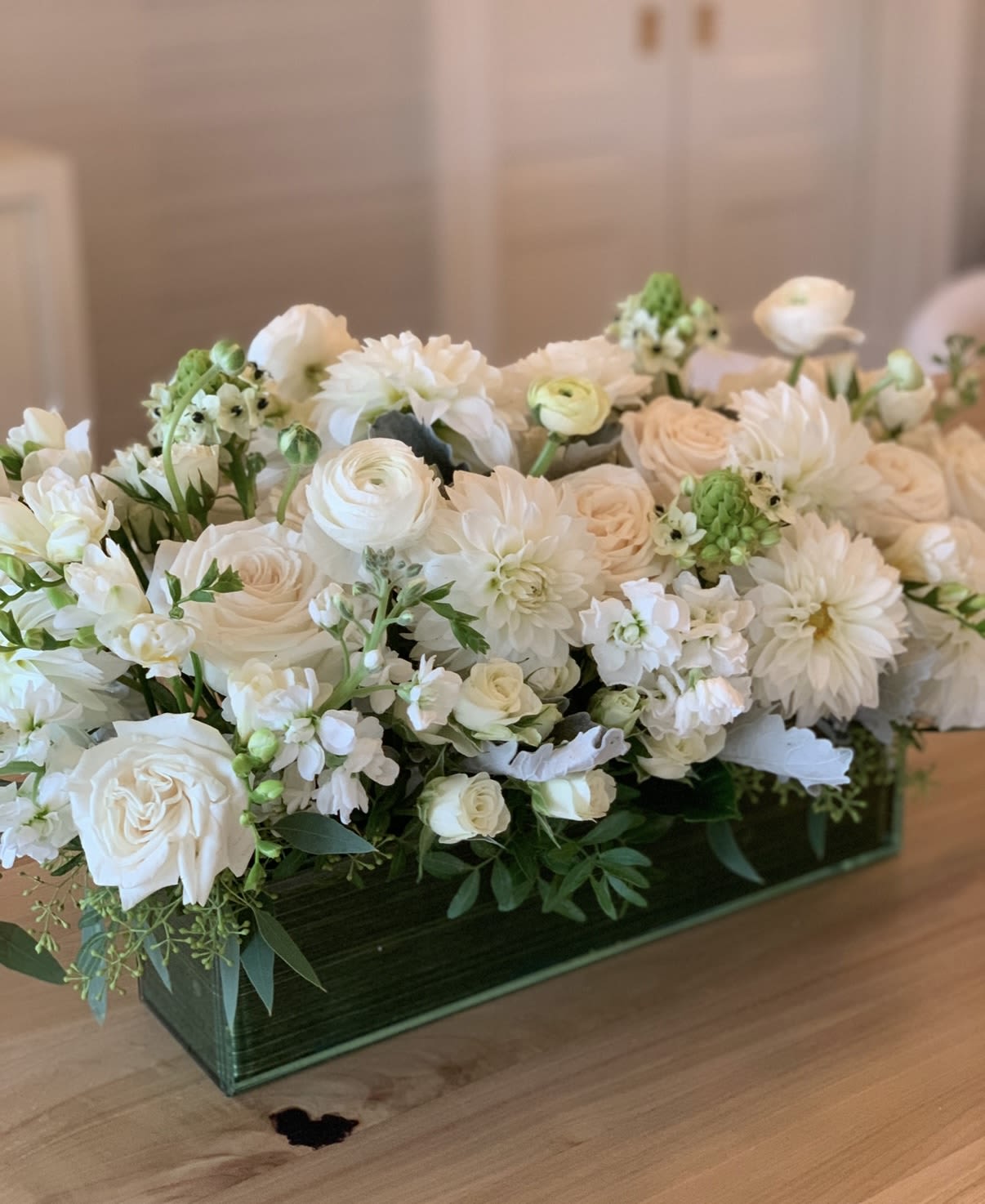 Mystical  - Closely packed white and cream-toned roses, dahlias and ranunculus sit amid sprays of stock for subtle height and scent, the textures blending from velvety petals to soft, layered blooms. The arrangement is presented in a sleek rectangular green glass vase that anchors the composition and highlights the neutral palette; its low, elongated shape is designed to run along dining tables or credenzas without blocking conversation.  Ideal for formal dinners, wedding guest tables, or as a thoughtful everyday gift, this arrangement brings an elegant, composed presence to any setting. Because of its low profile and dense packing, it's easy to place down the center of a table; keep blooms in fresh water and away from direct heat to prolong their life.