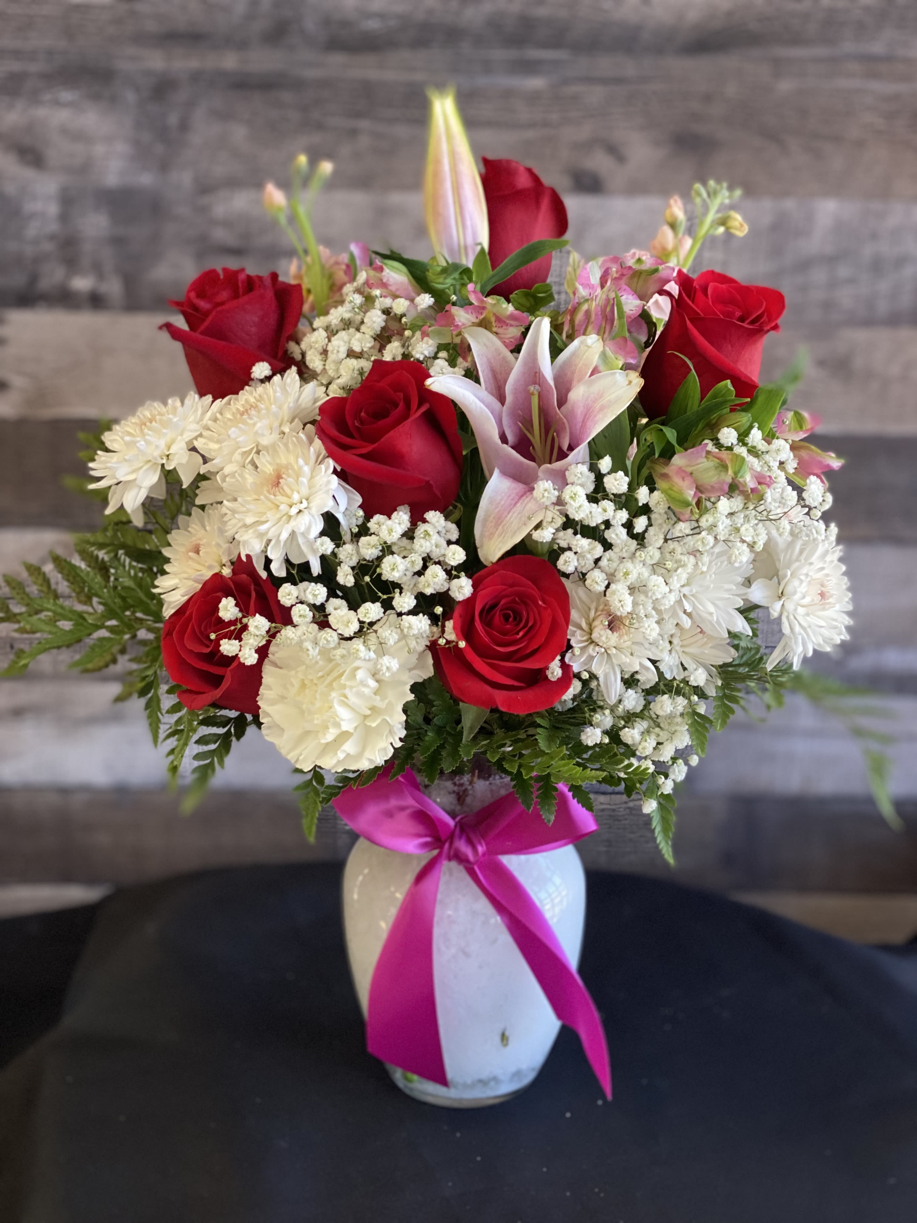 Bouquet of red roses, pink lilies, and white daisies in a white vase with a pink ribbon