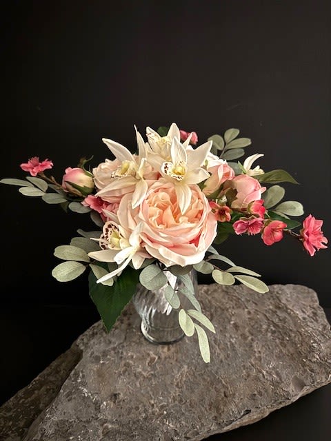 #A066 - Pink Cabbage Rose Silk Floral Bouquet - A single lush peach cabbage rose sits at the heart of this bridal bouquet, surrounded by creamy white cymbidium orchids and delicate pink spray roses-an elegant wedding bouquet that reads soft, romantic, and timeless. The asymmetrical, hand-tied shape and silvery-green foliage give it a slightly rustic, natural feel, perfect for a bride who wants fresh-from-the-garden charm for her ceremony or intimate reception. Crafted in high-quality silk, the bouquet features peach, white, pink, and green tones with orchid throats speckled for subtle visual interest. Stems are bound in textured white fabric ribbon and finished with a row of pearl buttons for a refined, bridal presentation.  The arrangement is shown gathered in a clear ribbed glass vase for centerpiece styling - compact and versatile, about 12" wide by 10.5" high.  It works equally well as a hand bouquet or a romantic table accent. Made to last beyond the day, this silk design lets the person you're celebrating keep a beautiful reminder of the moment. Ideal for weddings and anniversaries, it arrives ready to carry or display; light misting of dust and occasional gentle reshaping of stems will keep the colors and form looking fresh for years.