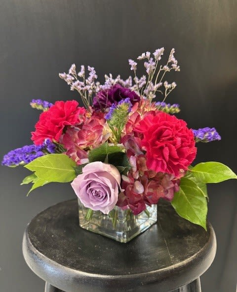 Low arrangement of pink and purple flowers with a lavender rose in a square glass vase.