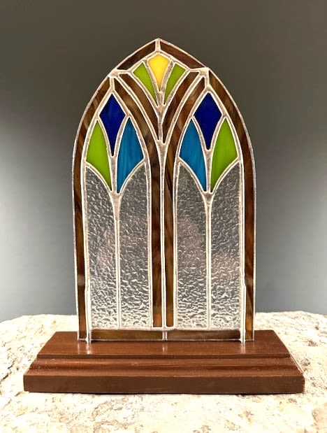 #A078 - Gothic Window Standing Sculpture - A small arched stained-glass panel standing on a tiered wood base creates a quietly soulful accent piece perfect for a sympathy keepsake, thinking-of-you gift, or a meaningful Mother's Day or anniversary offering. This window offers the reflective, chapel-like presence of blue, green and a single yellow jewel-toned pane on a small scale to deliver a contemplative, comforting impression-the kind that makes the recipient feel seen and remembered. The stained-glass window features clear textured lower panels framed by warm brown-toned strips and geometric, cathedral-inspired sections above; the piece sits upright on a sturdy wood base that reads like a miniature altar or mantel focal. Glasses in tones-soft blues, cool greens, and a hint of yellow add gentle texture without overwhelming the reflective design. Ideal for occasions from sympathy, get well, and thinking of you to birthdays, housewarmings, graduations, or holiday displays like Christmas and Hanukkah, this standing sculpture works year-round as a decorative keepsake. It's compact enough for a bedside table or entry shelf for a long-lasting memorial. Order options available for same-week delivery and personalization help ensure the piece arrives ready to comfort or celebrate the person you have in mind.