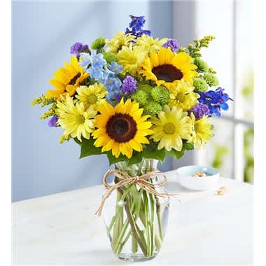 Fields Of Europe Summer - Inspired by the colorful charm of the European countryside, our best-selling summer bouquet is gathered with fresh-picked blooms, revealing the rustic beauty of nature. Shades of golden yellow and rich blue pop against lush greenery, creating a timeless gift for someone special to enjoy. • All-around arrangement with sunflowers, blue delphinium, yellow daisy poms, green button poms and purple statice, accented with assorted greenery • Artistically designed in a clear glass vase accented with raffia; measures 8""H • Extra-large arrangement measures approximately 19""H x 13.5""W • Large arrangement measures approximately 18""H x 13""W • Medium arrangement measures approximately 17""H x 11.5""W • Our florists hand-design each arrangement, so colors, varieties and container may vary due to local availability • Sunflowers may arrive with light or dark centers"