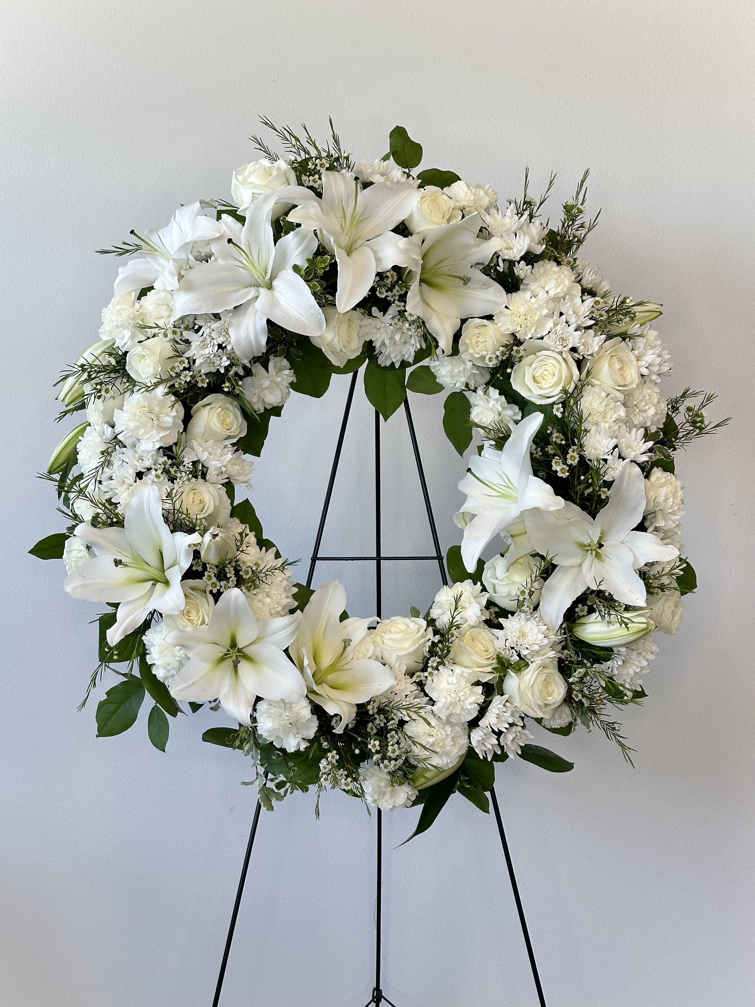 Serenity Wreath - A gentle embrace for tender hearts, the Serenity Wreath from Irvine Village Flowers offers a tranquil ring of fragrant white blossoms, a quiet tribute for any funeral or wake. Delivered on an easel, this classic arrangement whispers sympathy and reverence, crafted with care from fresh roses, Asiatic lilies, carnations, and cushion spray chrysanthemums, each bloom cradled by soft greenery. Measuring 22" W x 22" H, its one-sided grace stands as a thoughtful beacon of peace.    The pure white roses speak of honor, remembrance, and quiet love, their petals a tender solace. Asiatic lilies, radiant and calm, bloom with dignity and hope, easing sorrow with their gentle presence. Carnations offer enduring affection and resilience, while cushion spray chrysanthemums lend a comforting fullness, symbolizing truth and eternal rest. Together, they weave a wreath of serenity, a heartfelt gesture for those in mourning.    In this time of stillness, Irvine Village Flowers is here for you—visit us online or in-store to share this compassionate creation. With our seamless delivery across Irvine, your tribute arrives fresh, a delicate balm to honor and console.