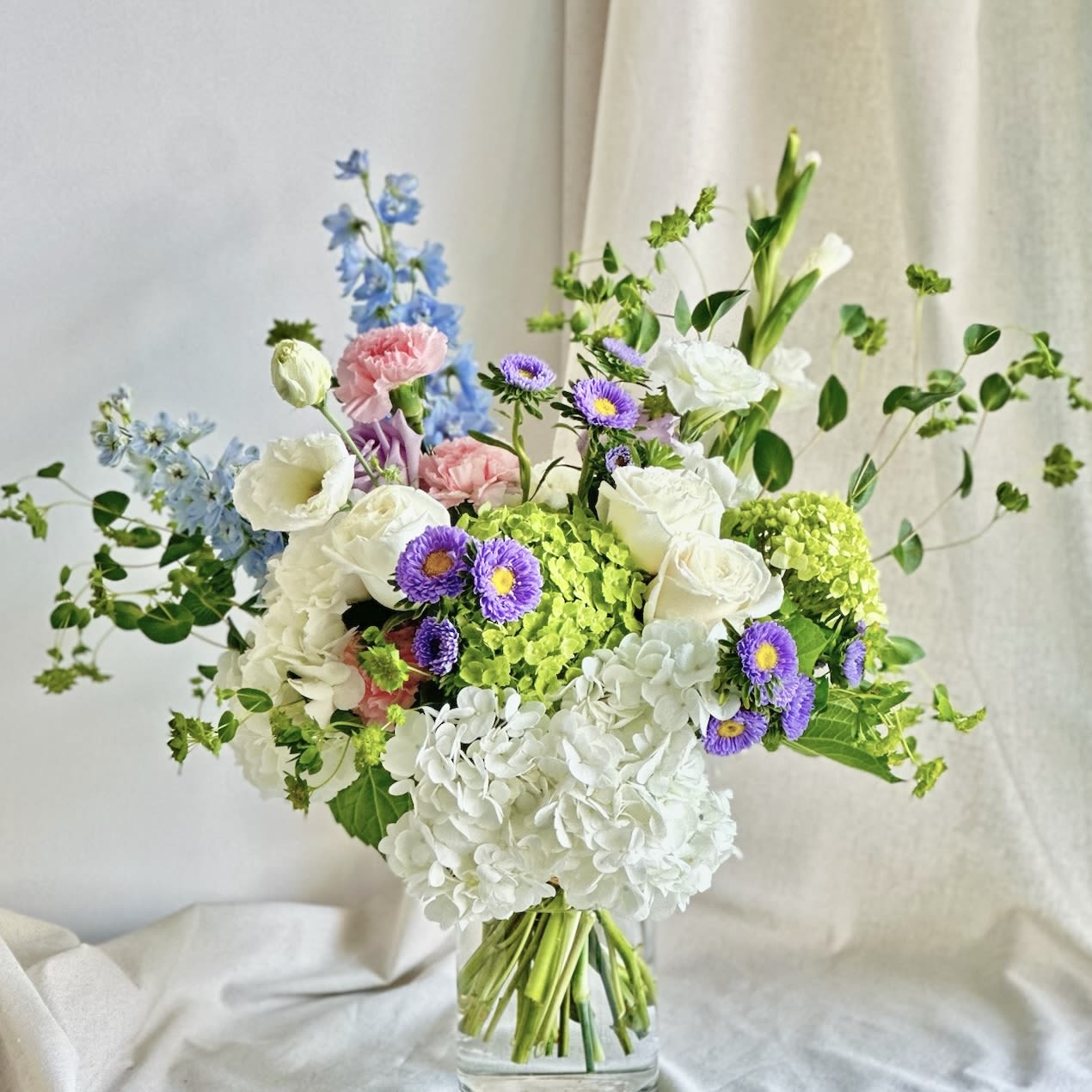 Sparks Fly  - A medium cylinder glass vase with white, lavender, pink, blue, and green flowers, arranged front-facing.   Flower types may vary depending on the season.