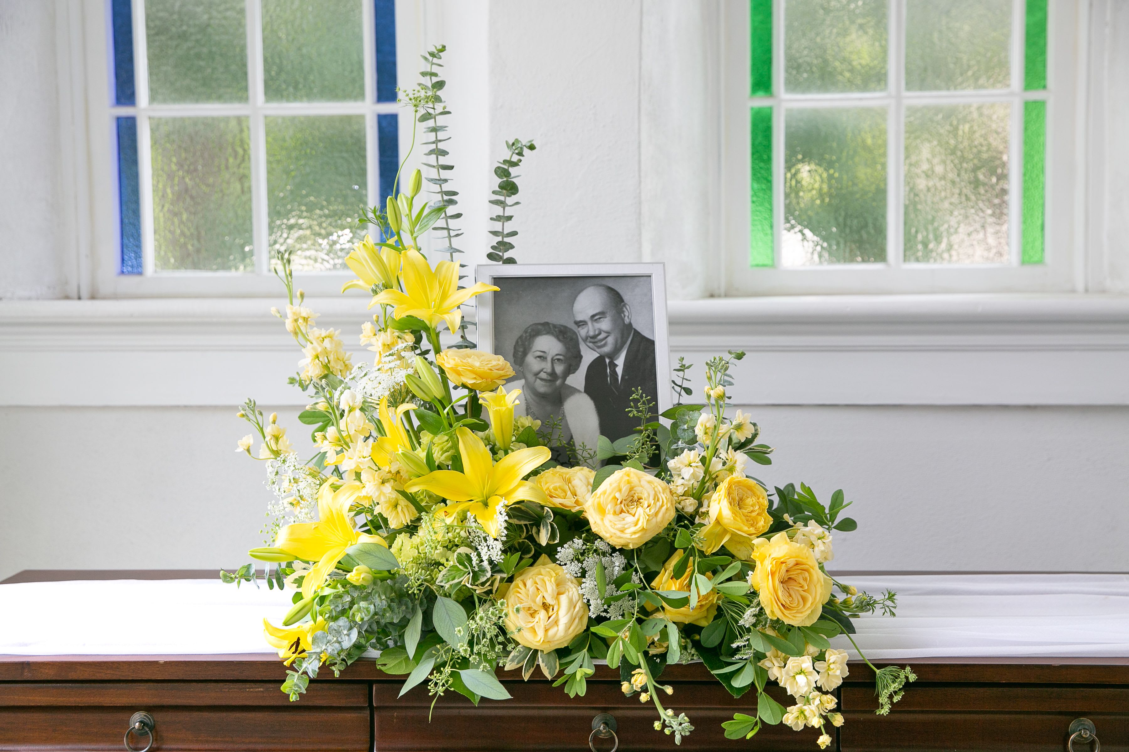 Yellow lily and rose sympathy arrangement displayed with a framed black-and-white portrait.