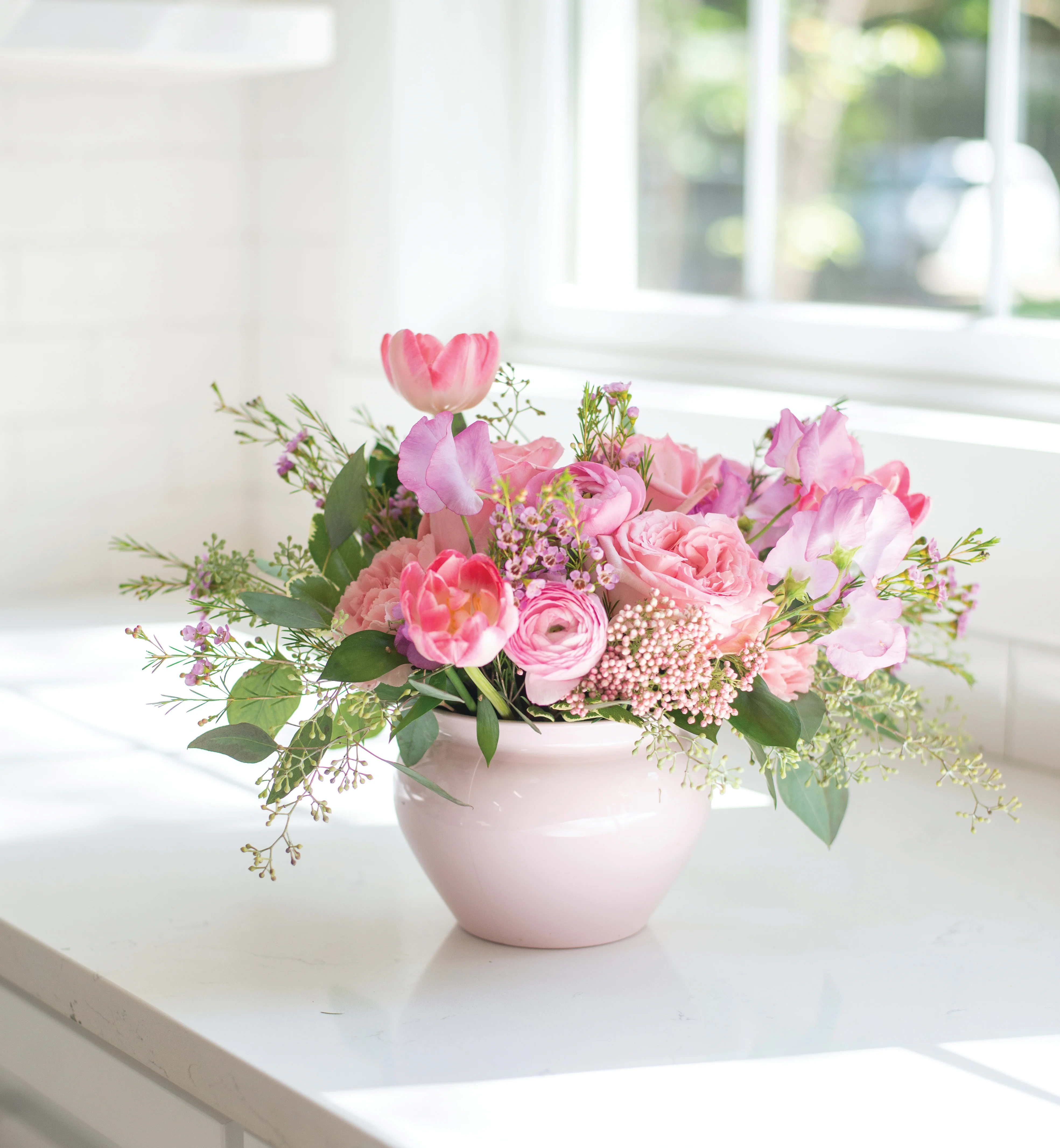 Low pink arrangement of tulips, roses, and ranunculus in a pale pink ceramic bowl