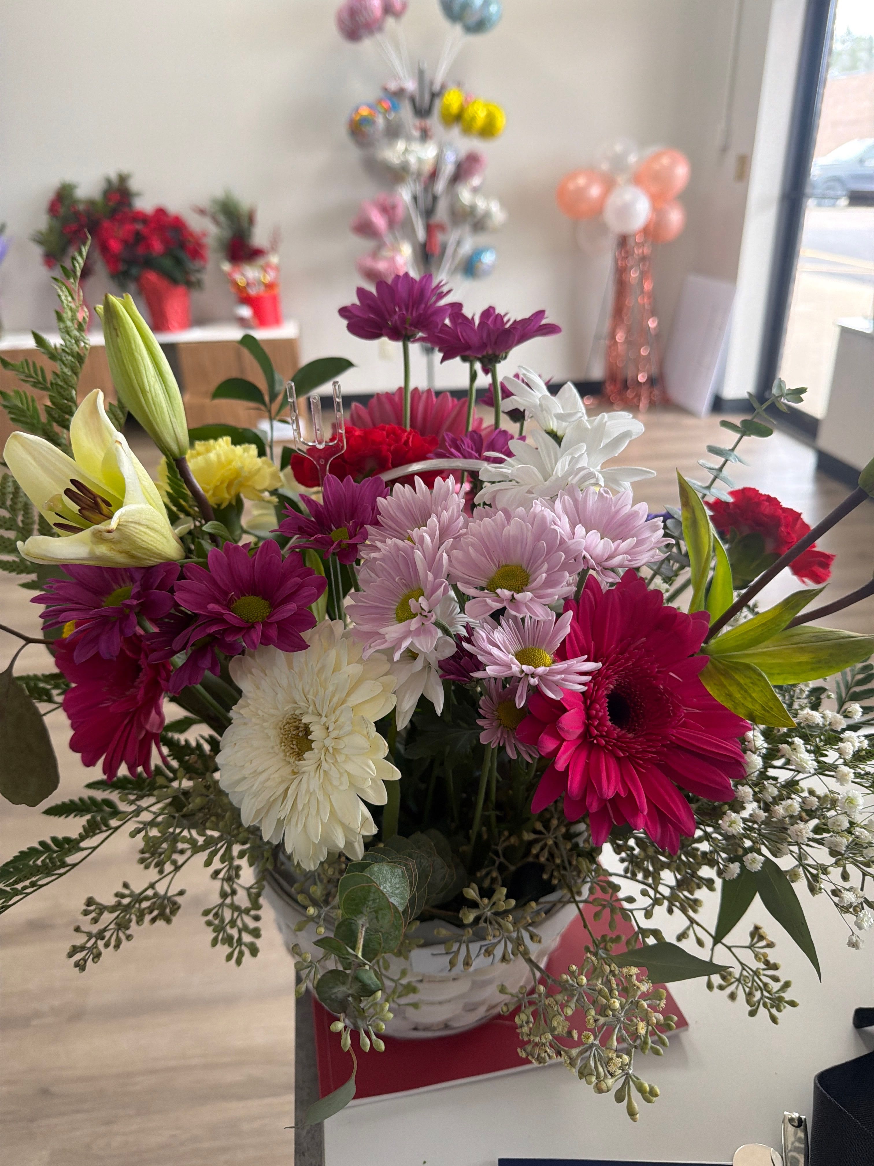 Cheerful  color burst -  A vibrant mix of fresh blooms designed to brighten any day. This arrangement features a lively assortment of gerbera daisies, lilies, chrysanthemums, carnations, and accent greenery, beautifully arranged in a decorative container. With its rich pinks, soft pastels, bright whites, and pops of yellow, this bouquet brings a cheerful, garden-fresh look to any space. Perfect for birthdays, celebrations, get-well wishes, or simply adding color and joy to someone’s home