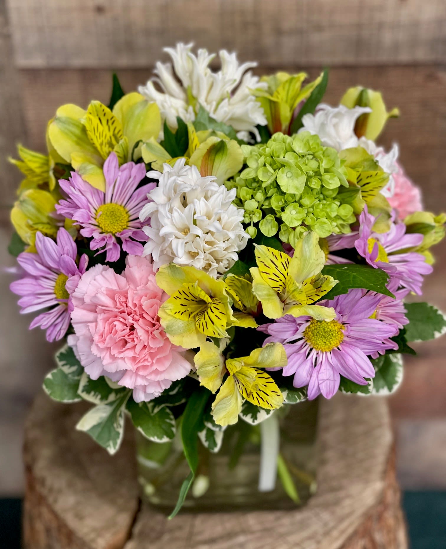 Spring Awakening - A compact design of fresh pastels, this spring flower arrangement is a cheerful pick-me-up or birthday bouquet that brightens a desk, dining table, or bedside. Yellow alstroemeria with its dark speckling threads through clusters of soft pink carnations and lavender daisy chrysanthemums, while a single bright green hydrangea and white hyacinth add lush texture and a gentle scent that feels like the first warm day of the season.